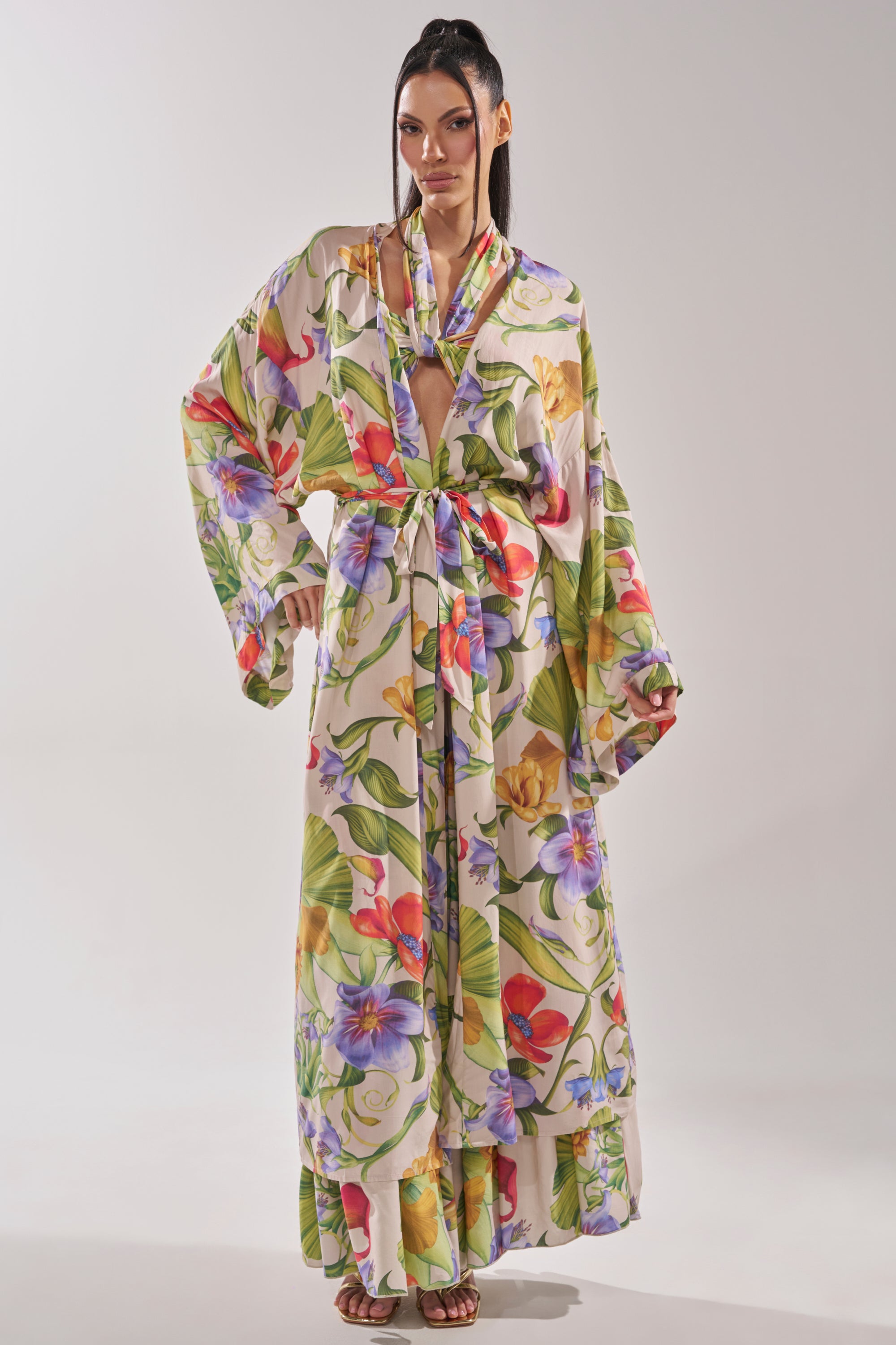 A woman with straight, dark hair poses against a light background wearing the INTO THE GARDEN CARDIGAN, a long, flowing lightweight cover-up with a colorful floral print, wide sleeves, deep V-neck, and tie belt at the waist. 0