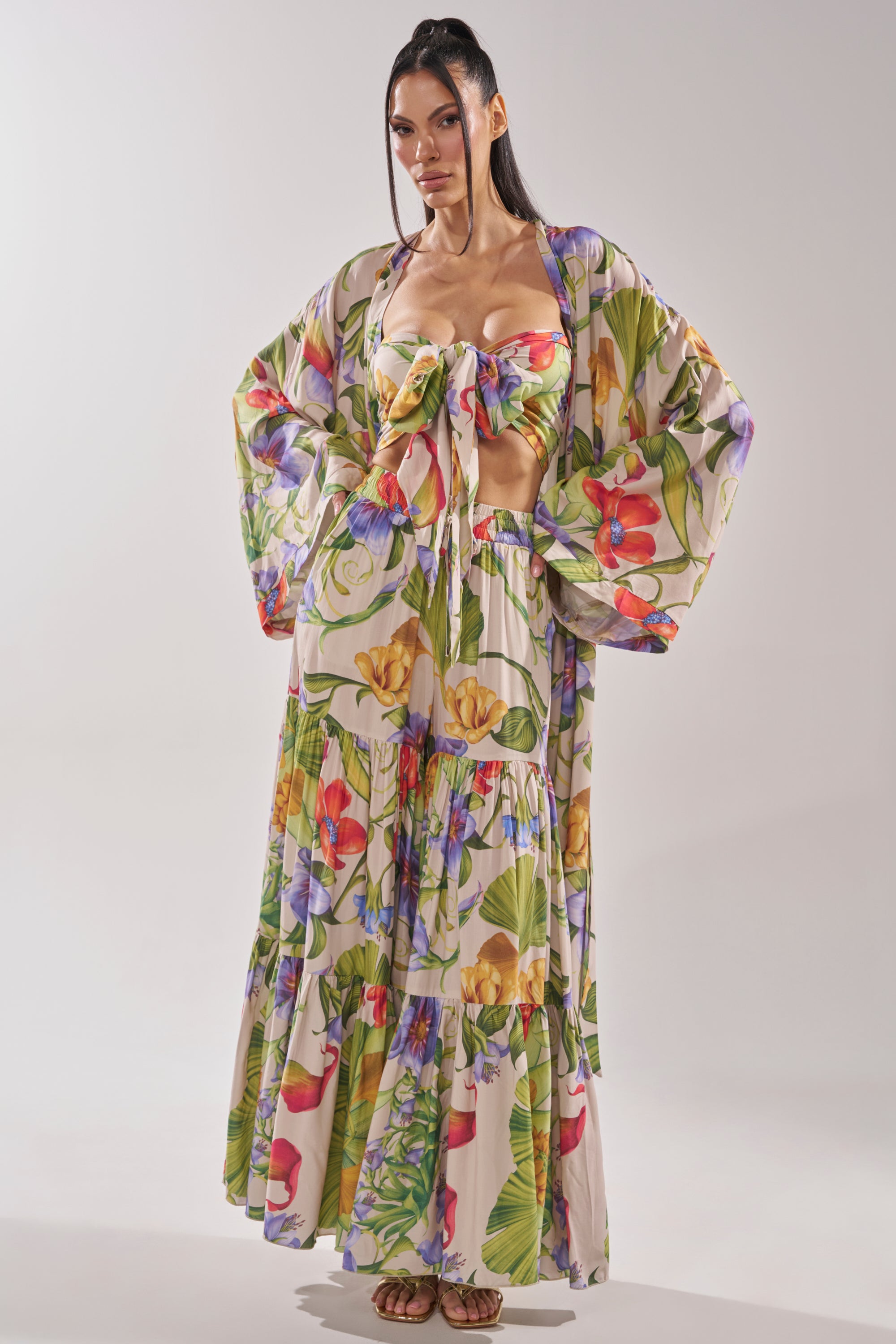 A woman stands confidently in the INTO THE GARDEN CARDIGAN, a flowy, long-sleeved tropical floral print robe, paired with a matching tie-front crop top and tiered maxi skirt. Her long dark hair is styled in two ponytails. 1