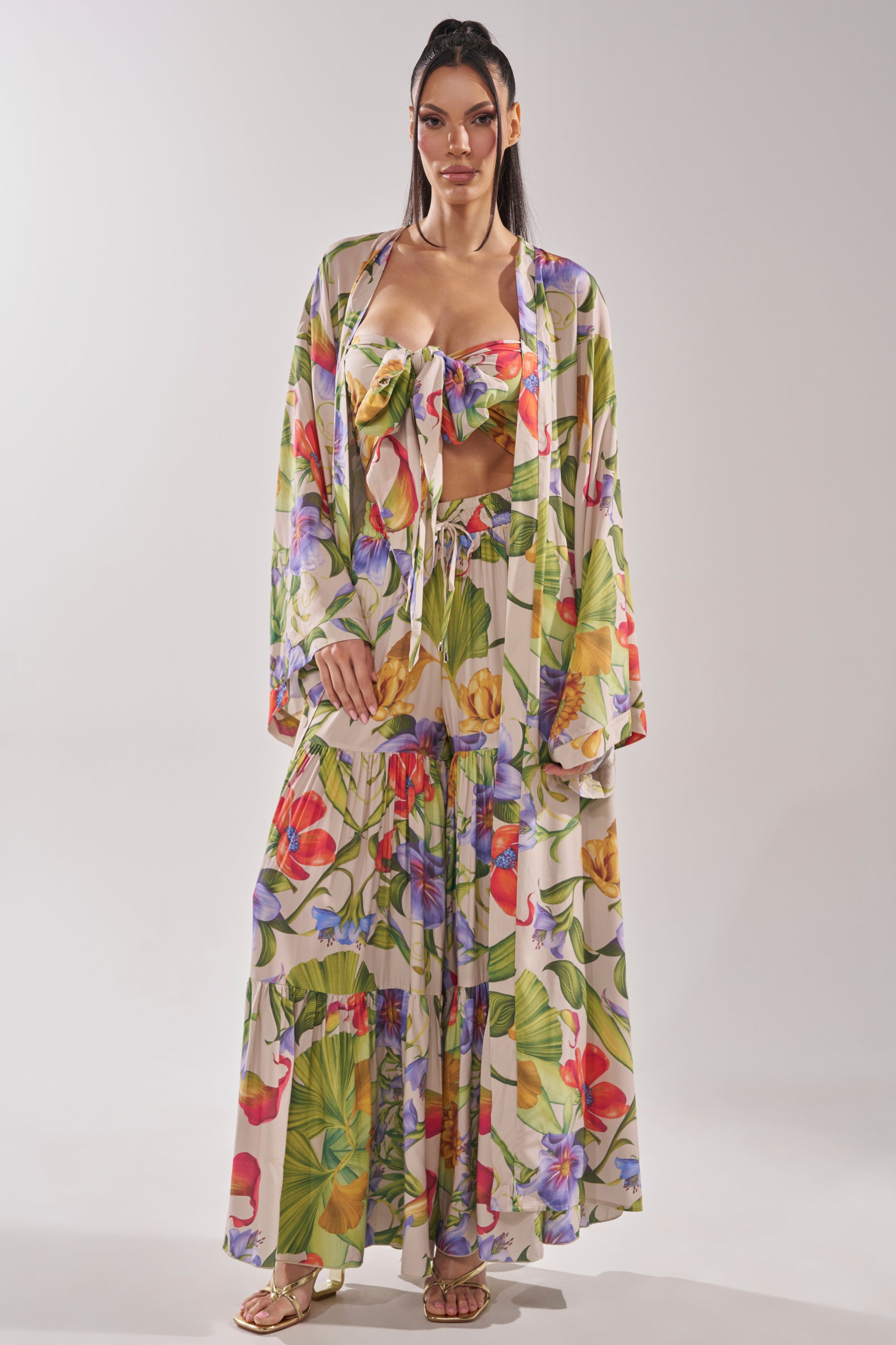 A woman wears the INTO THE GARDEN CARDIGAN in a longline silhouette with a matching tropical floral bandeau and flowing skirt, styled with gold sandals against a plain, light background. 2