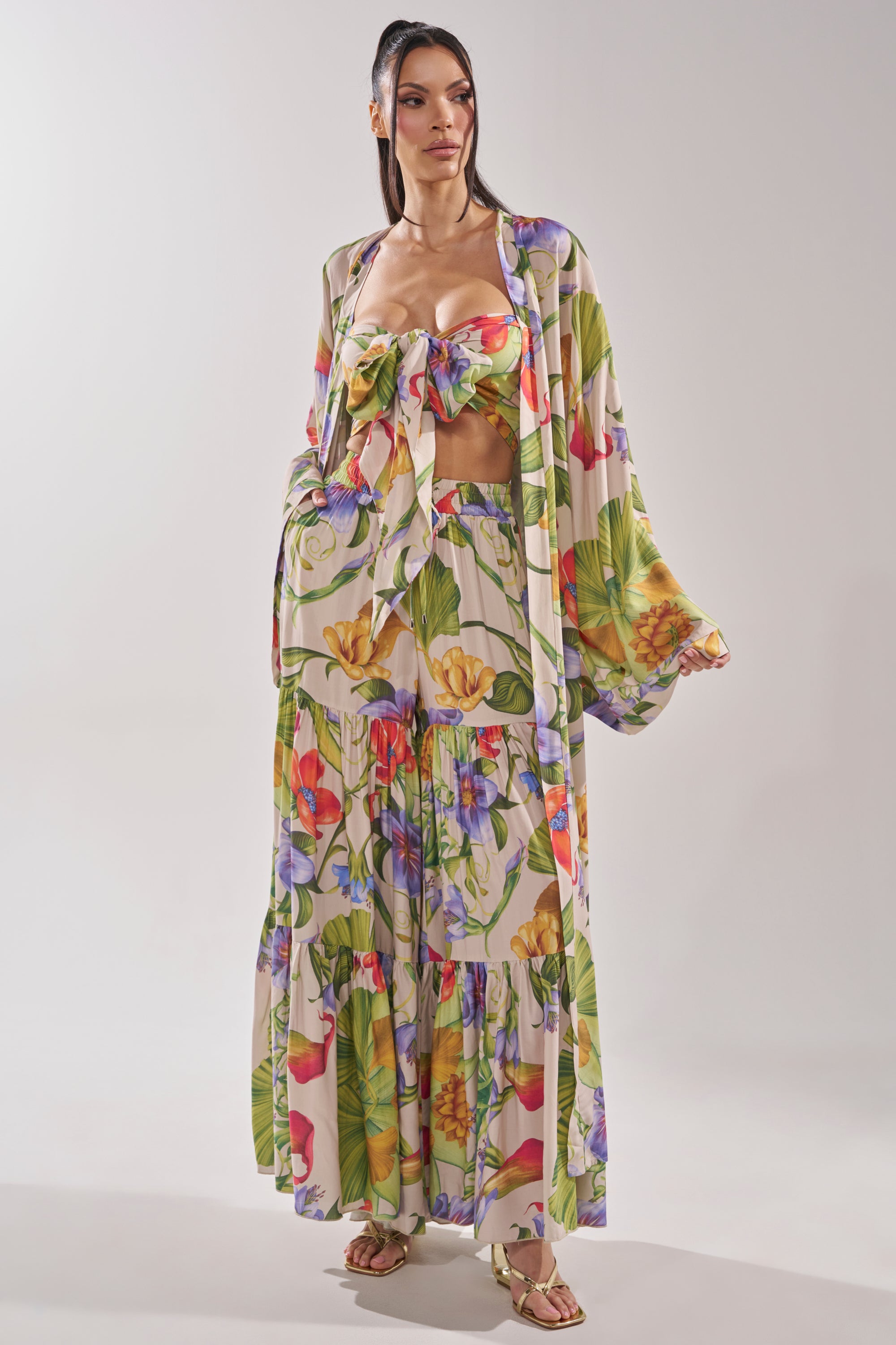 A woman models the INTO THE GARDEN CARDIGAN—a vibrant, tropical floral print longline kimono—paired with a matching bandeau top, wide-leg pants, and gold sandals against a plain background. 3