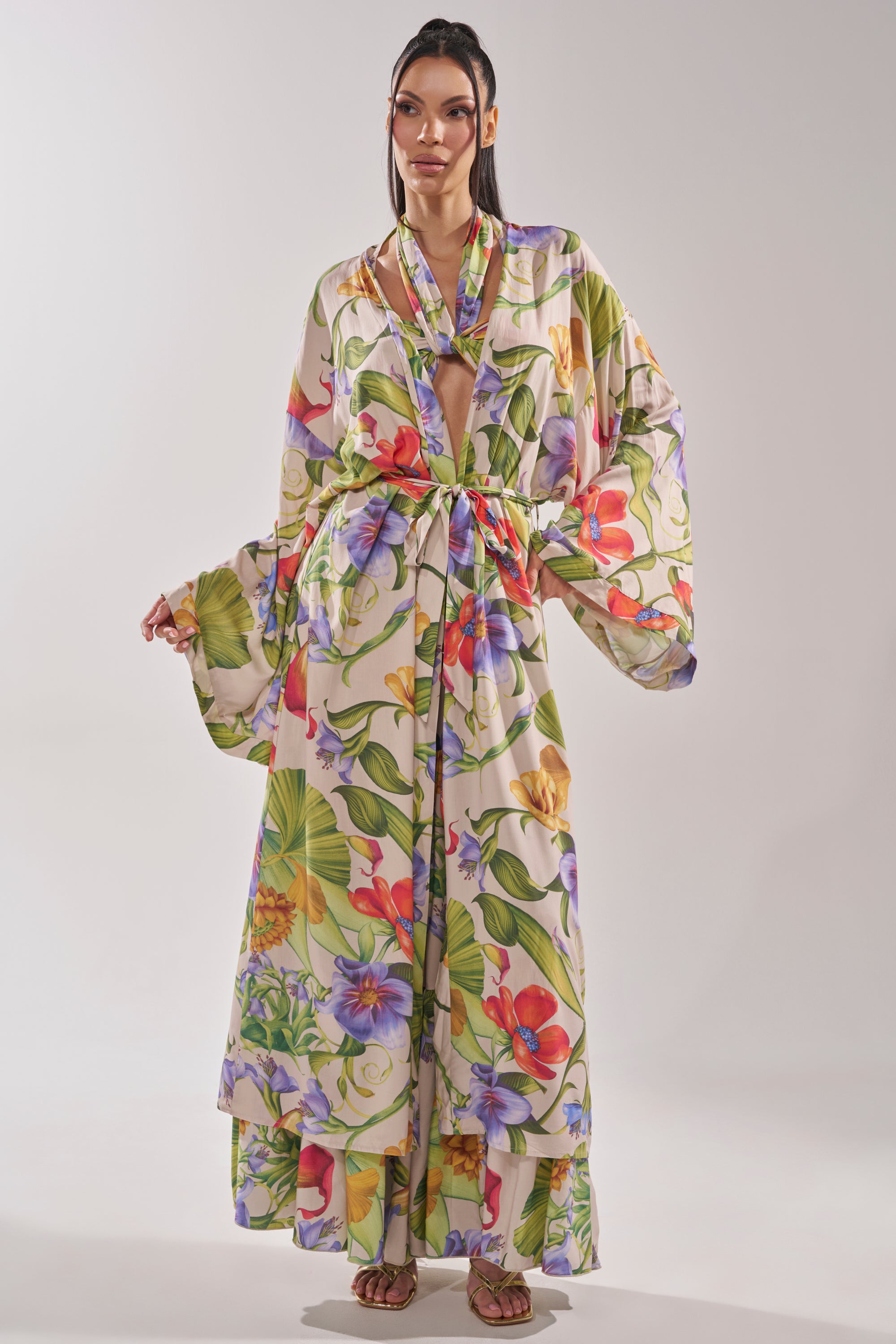 A woman models the INTO THE GARDEN CARDIGAN, featuring a tropical floral print, wide sleeves, a deep V-neck, and a belted waist. She wears sandals and poses against a simple light background. 4