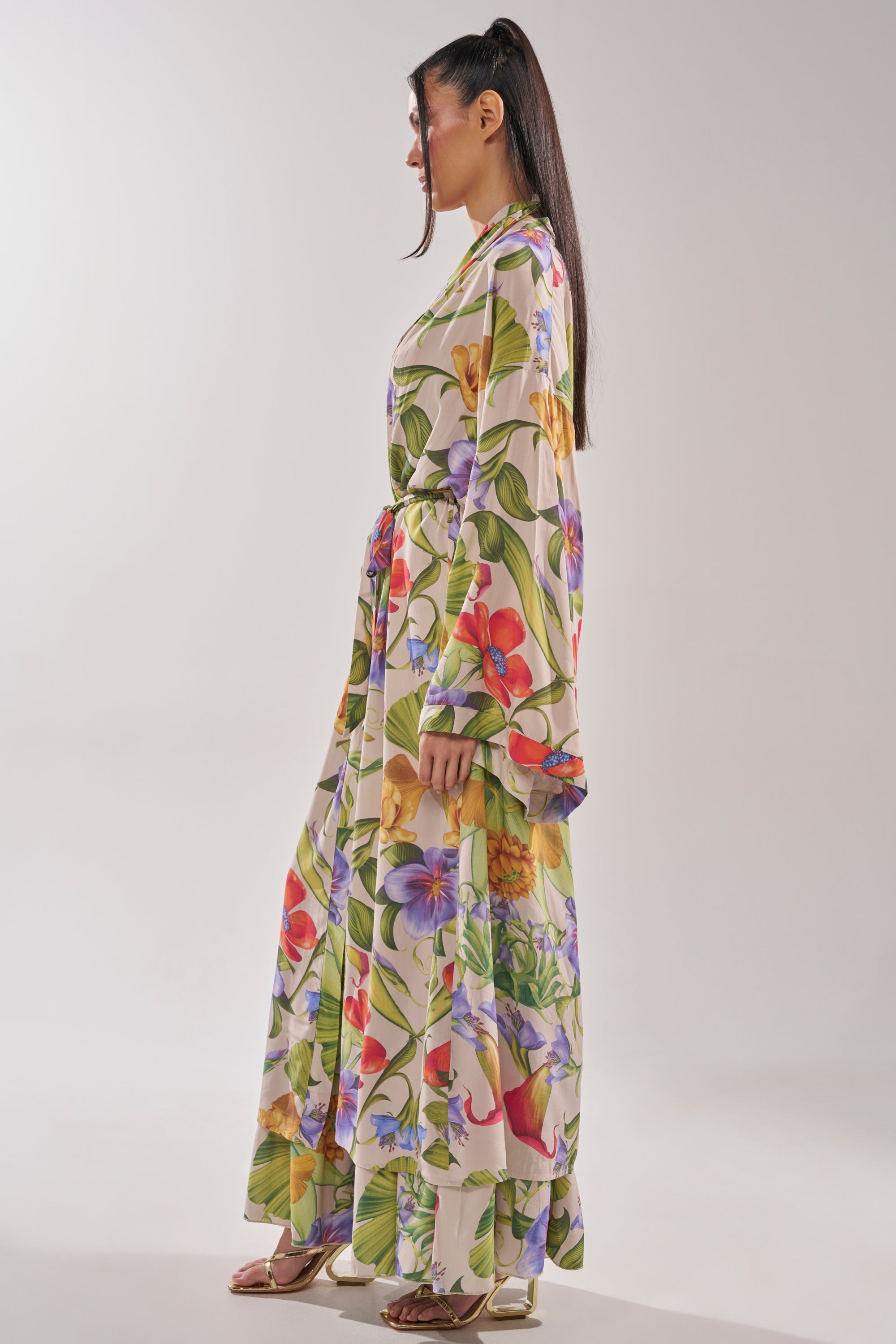 A woman in profile wears the INTO THE GARDEN CARDIGAN, featuring a vibrant tropical floral print, long sleeves, and a tie waist. Her sleek ponytail and sandals complete the look against a plain light gray background. 5