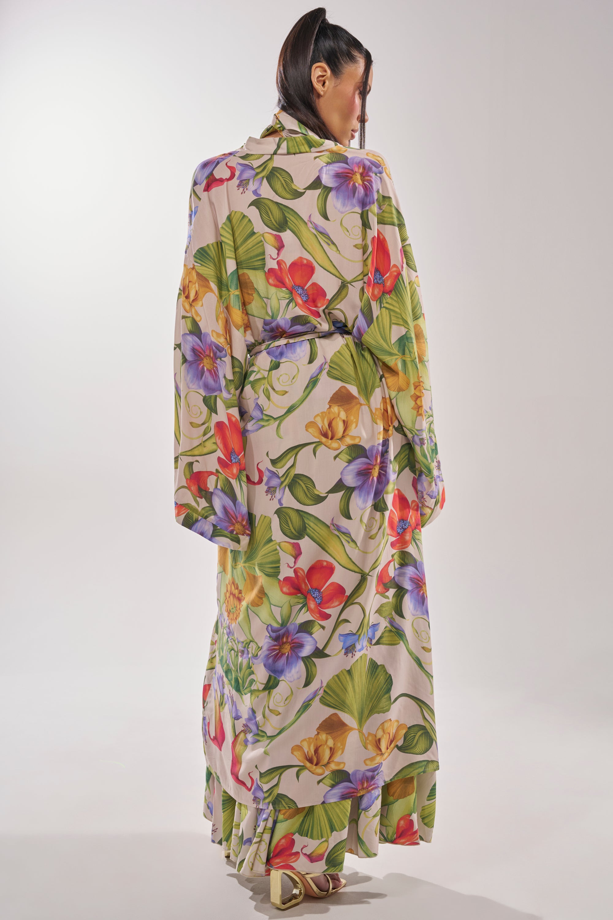 A woman faces away, wearing the INTO THE GARDEN CARDIGAN—a longline piece with a vibrant floral print. Her dark hair is tucked behind her ear as she stands against a plain, light background. 6