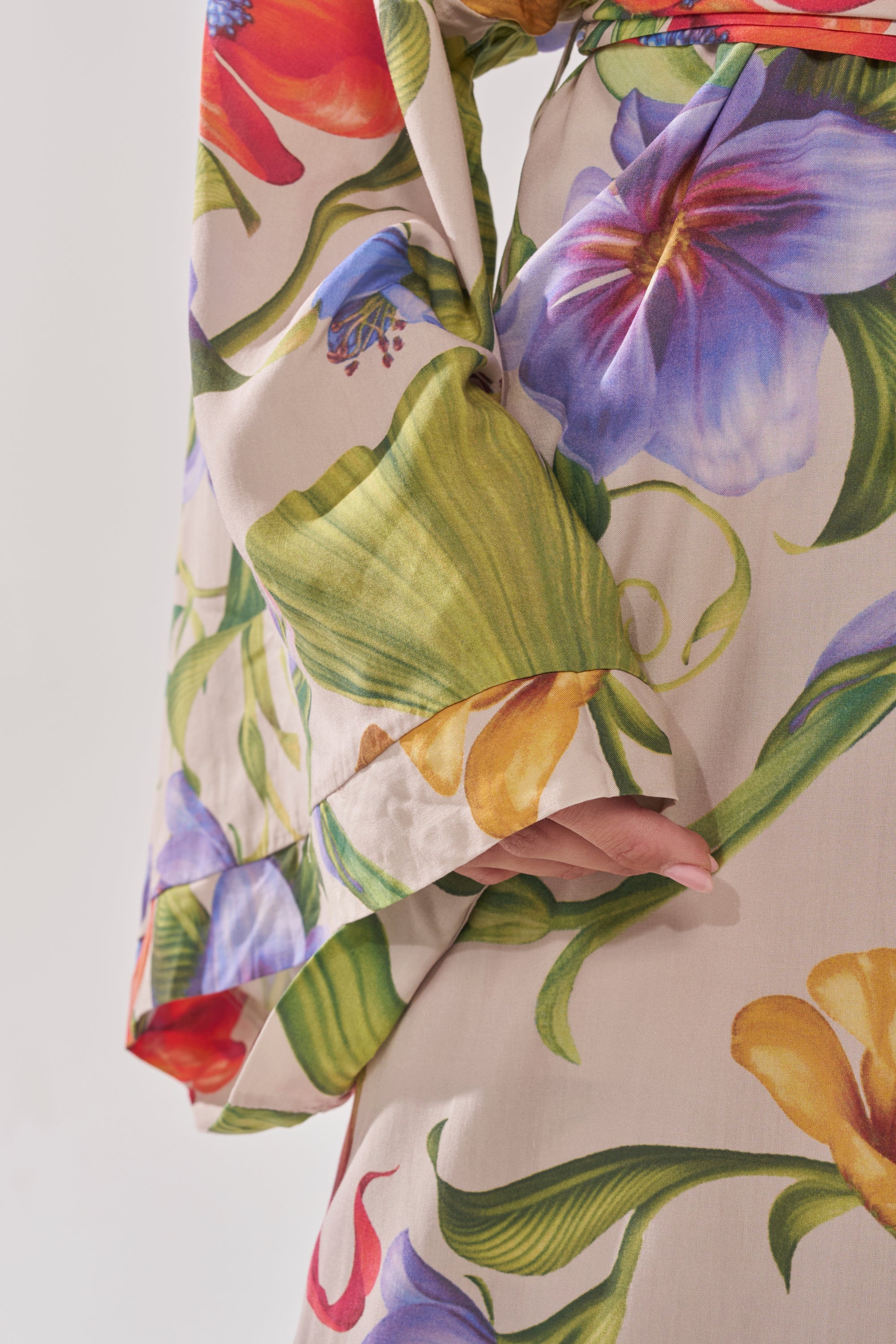 A close-up of a person’s arm and hand wearing the INTO THE GARDEN CARDIGAN, featuring a colorful floral print with bold leaves and flowers in purple, yellow, red, and green. 8