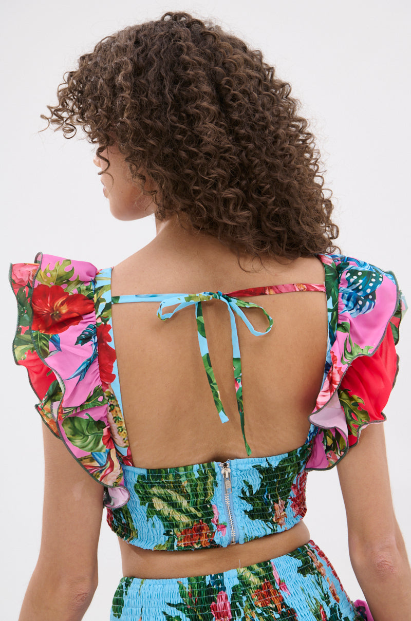 A person with curly hair wears the SOMETHING FEELS RIGHT FLORAL BLOUSE, a vibrant cropped top with ruffled sleeves and an open back tied with thin straps. The photo from behind against a plain background highlights its playful style.