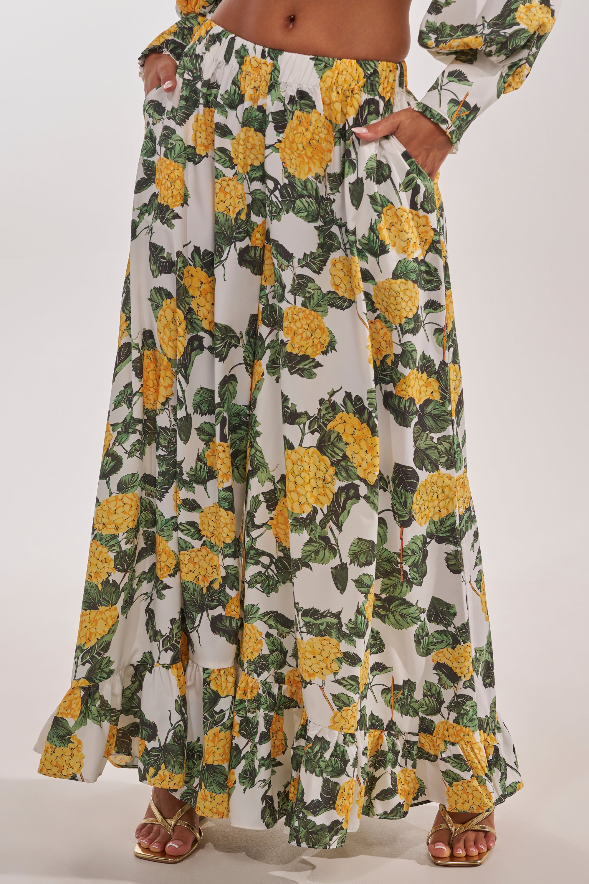 A person styles the JULIE FLORAL WIDE LEG RUFFLE PANT, featuring yellow florals and green leaves, ruffle hem, and pockets—perfect for vacation. The chic look is finished with gold sandals. 1