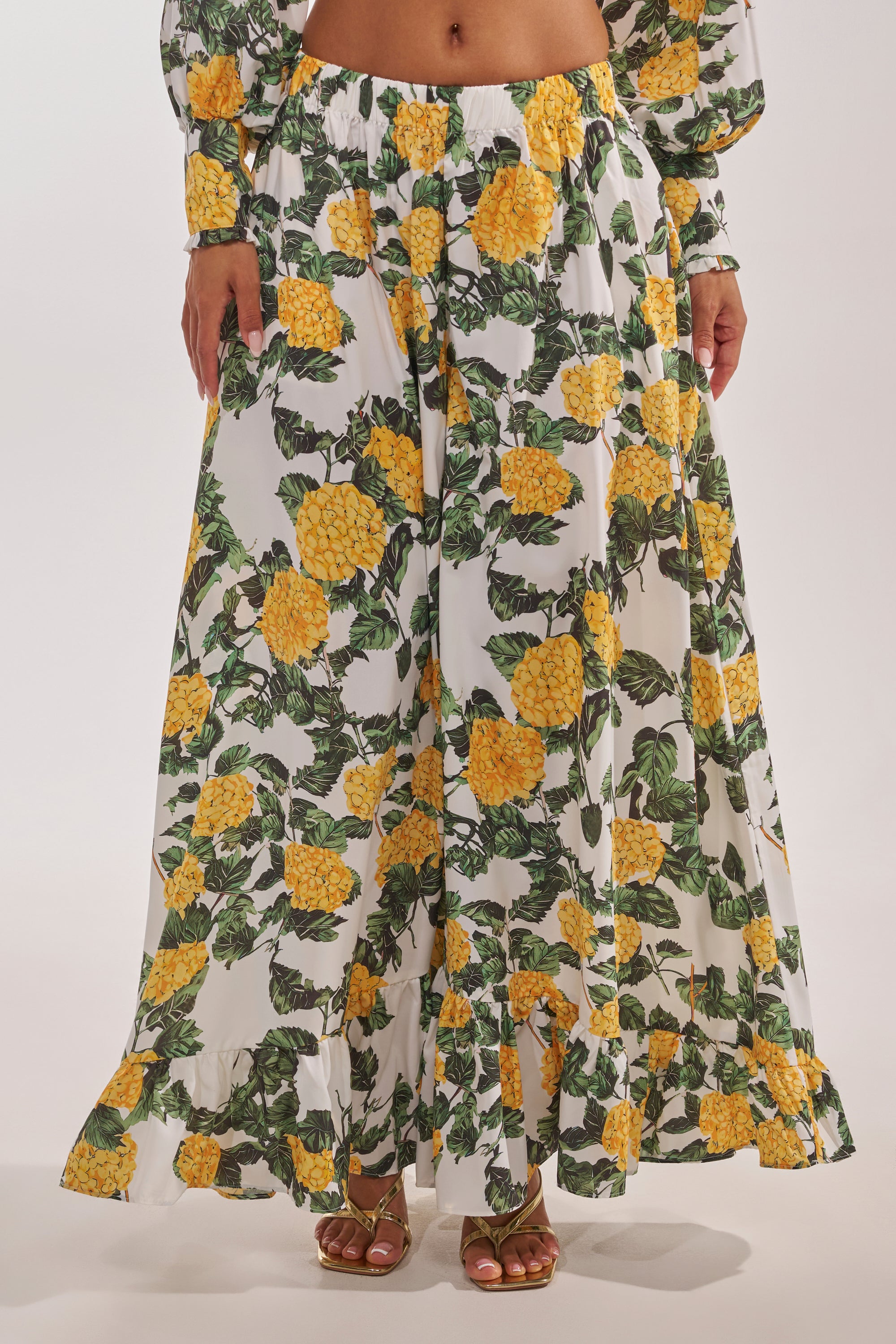 A person in JULIE FLORAL WIDE LEG RUFFLE PANT, featuring bright yellow florals and green leaves, styled with brown sandals—an effortlessly chic vacation look. Only the lower half of the body is visible. 2