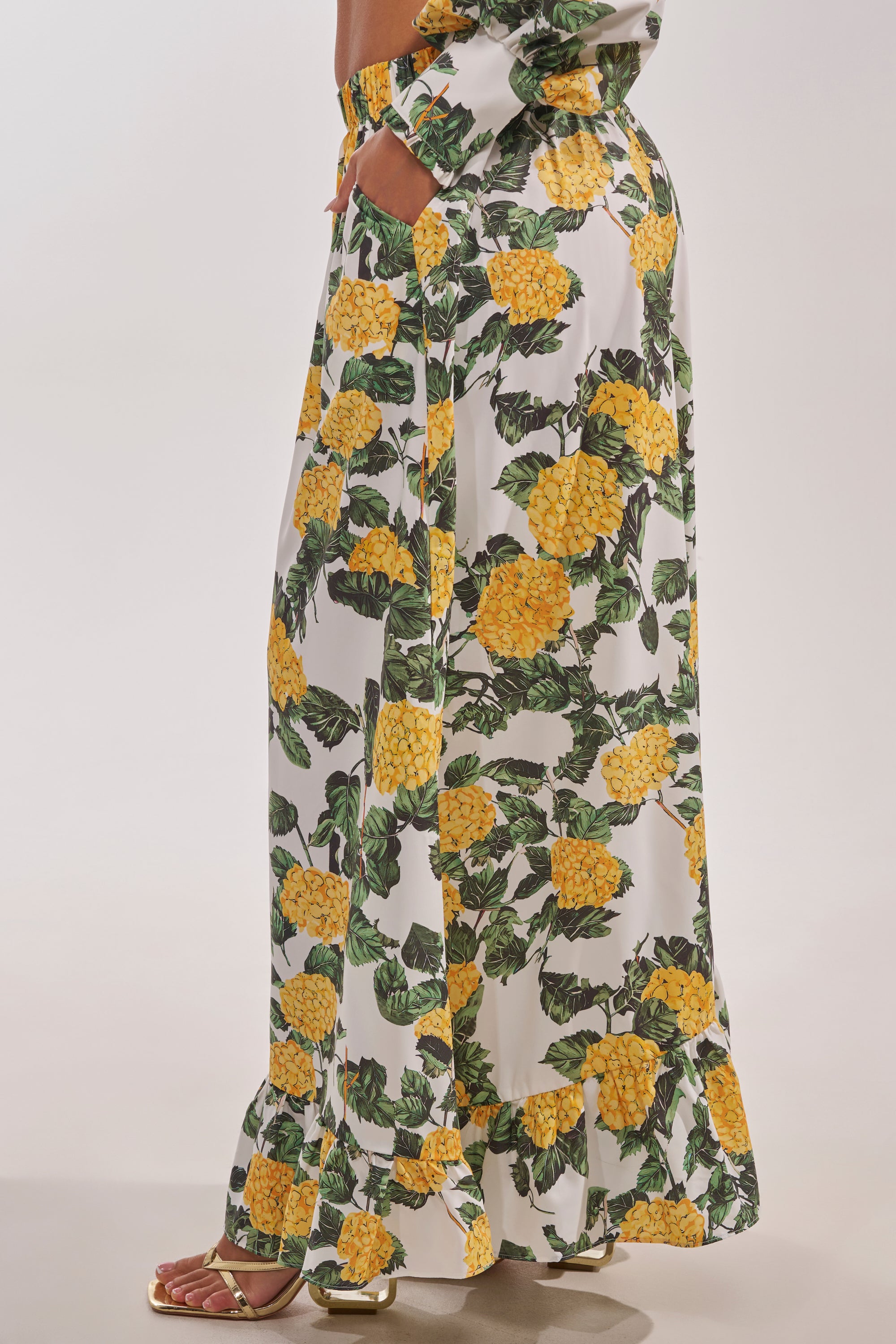A woman wears the JULIE FLORAL WIDE LEG RUFFLE PANT, featuring a white base with yellow floral print and ruffled hem—perfect for vacation—paired stylishly with metallic open-toe sandals. Only her lower body is shown. 3