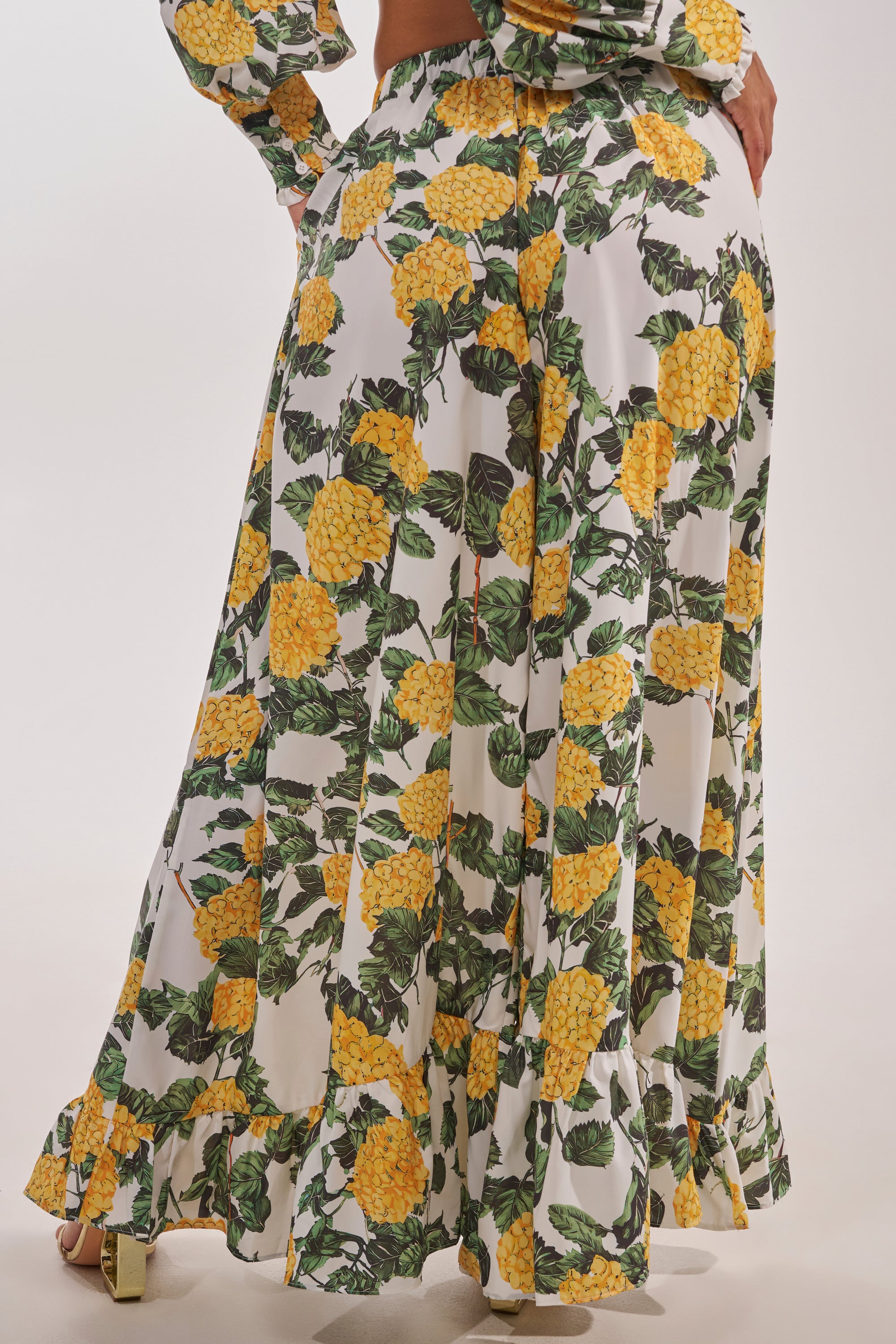 A close-up of a person wearing the JULIE FLORAL WIDE LEG RUFFLE PANT, featuring a vibrant yellow pineapple and green leaf print with flowy white fabric, styled with gold heels—ideal for vacation looks. 4