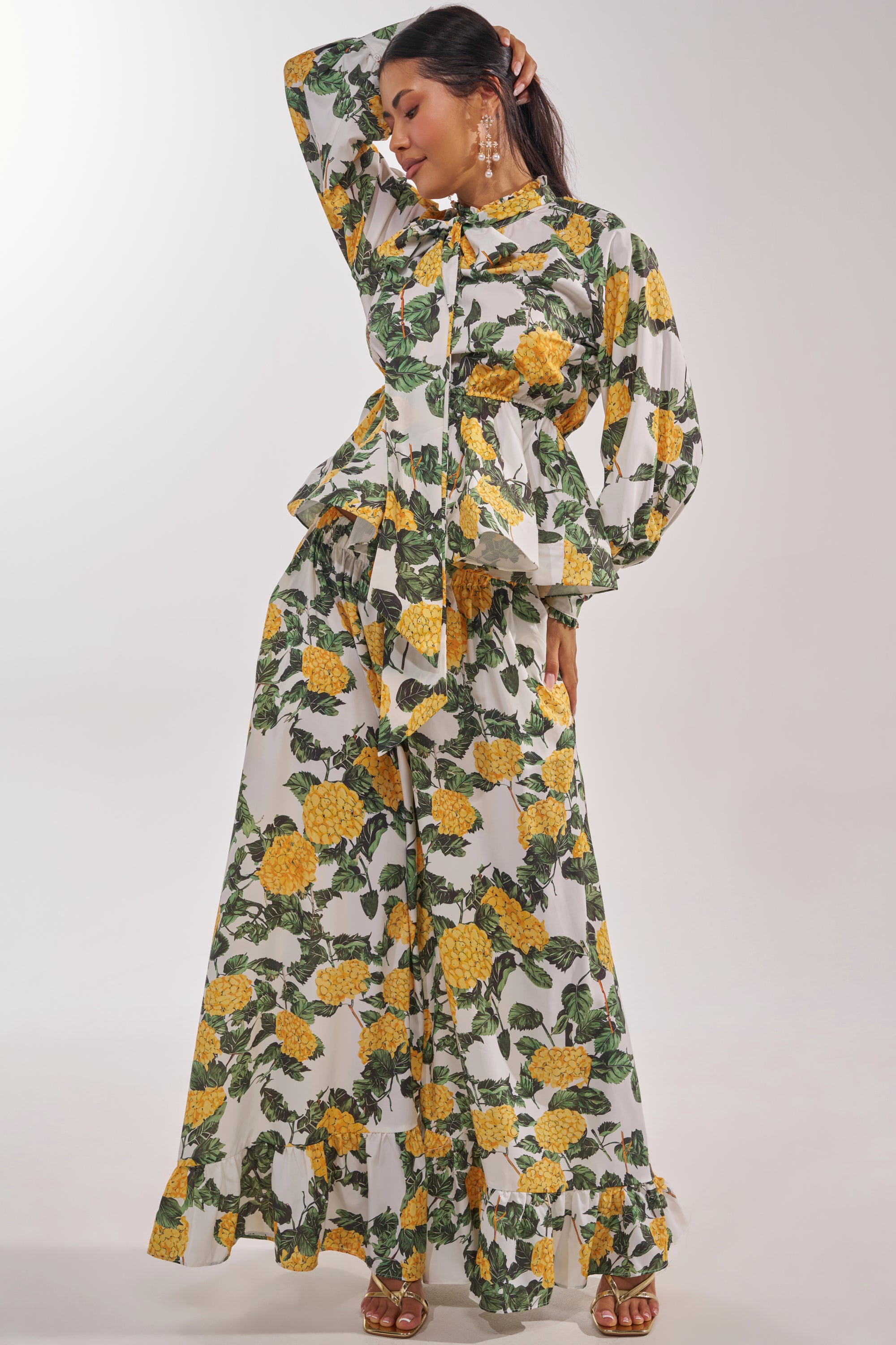 A woman models the JULIE FLORAL WIDE LEG RUFFLE PANT, featuring a vibrant yellow floral print and green leaves. She completes her vacation-ready look with gold earrings and heeled sandals, exuding effortless style. 5