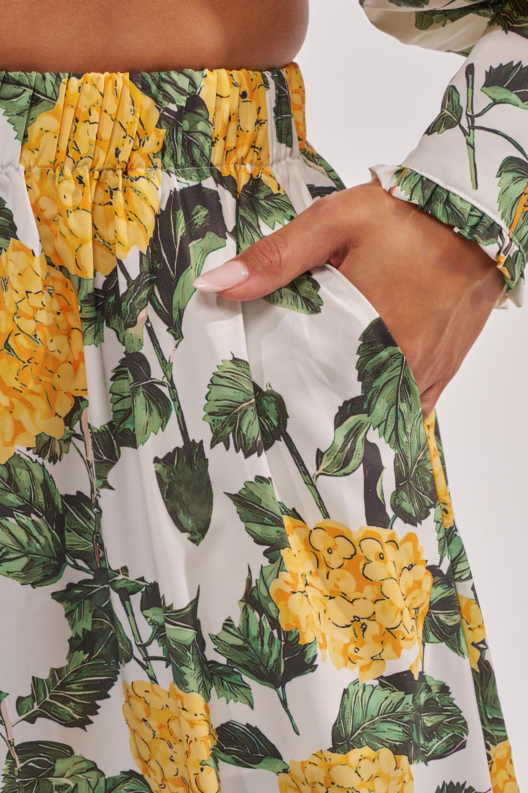 A close-up shows someone wearing the JULIE FLORAL WIDE LEG RUFFLE PANT, featuring yellow fabric with green leaf prints. Their right hand is in the pocket and the sleeve displays a matching floral design—perfect for vacation style. 6