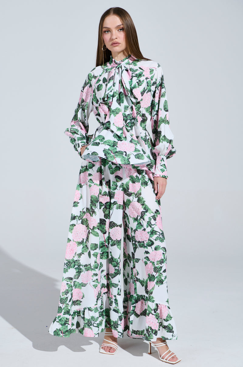 A woman poses against a neutral backdrop wearing the Diane Floral Wide Leg Ruffle Pant, featuring green, pink, and white patterns. She complements the look with white strappy heels and her long hair cascading straight as she gazes forward, embodying effortless elegance.