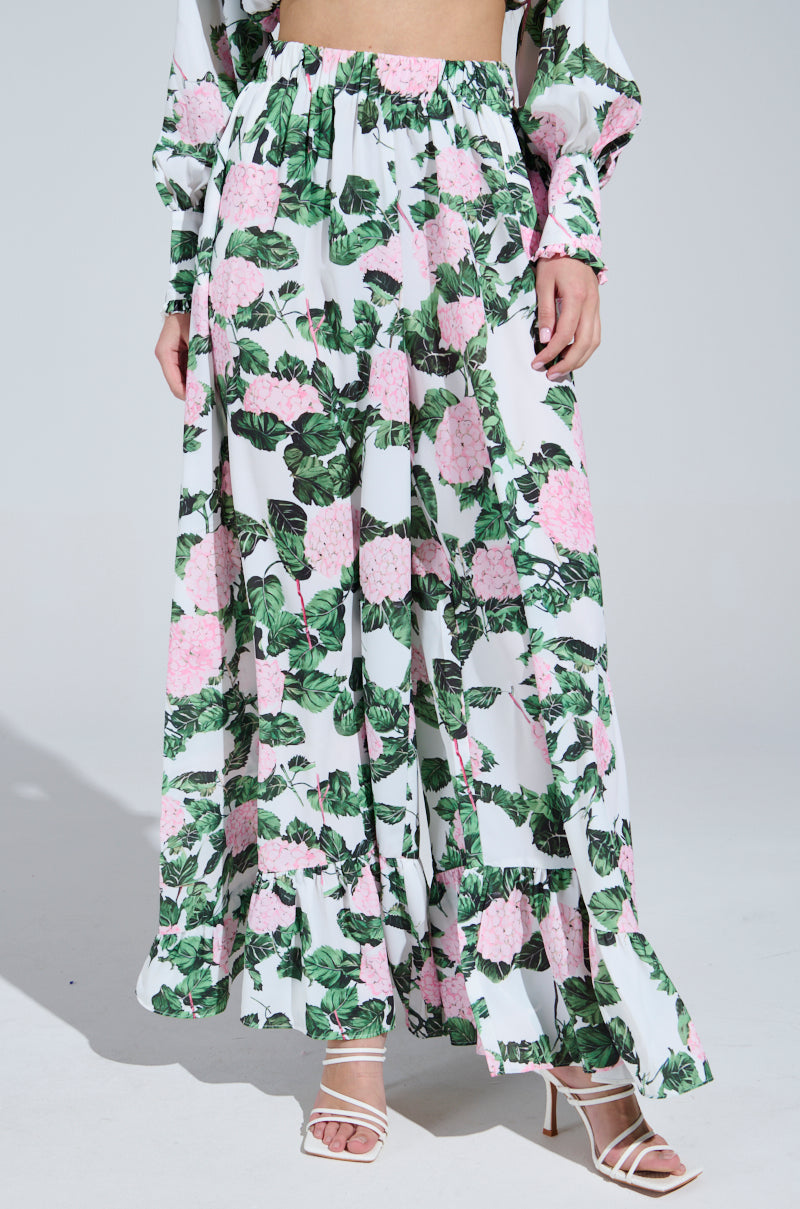 A person wearing a DIANE FLORAL satin set with pink flowers and green leaves, featuring a long-sleeved top and matching wide-leg ruffle pants. They're paired with white strappy high-heeled sandals, standing against a plain gray background.