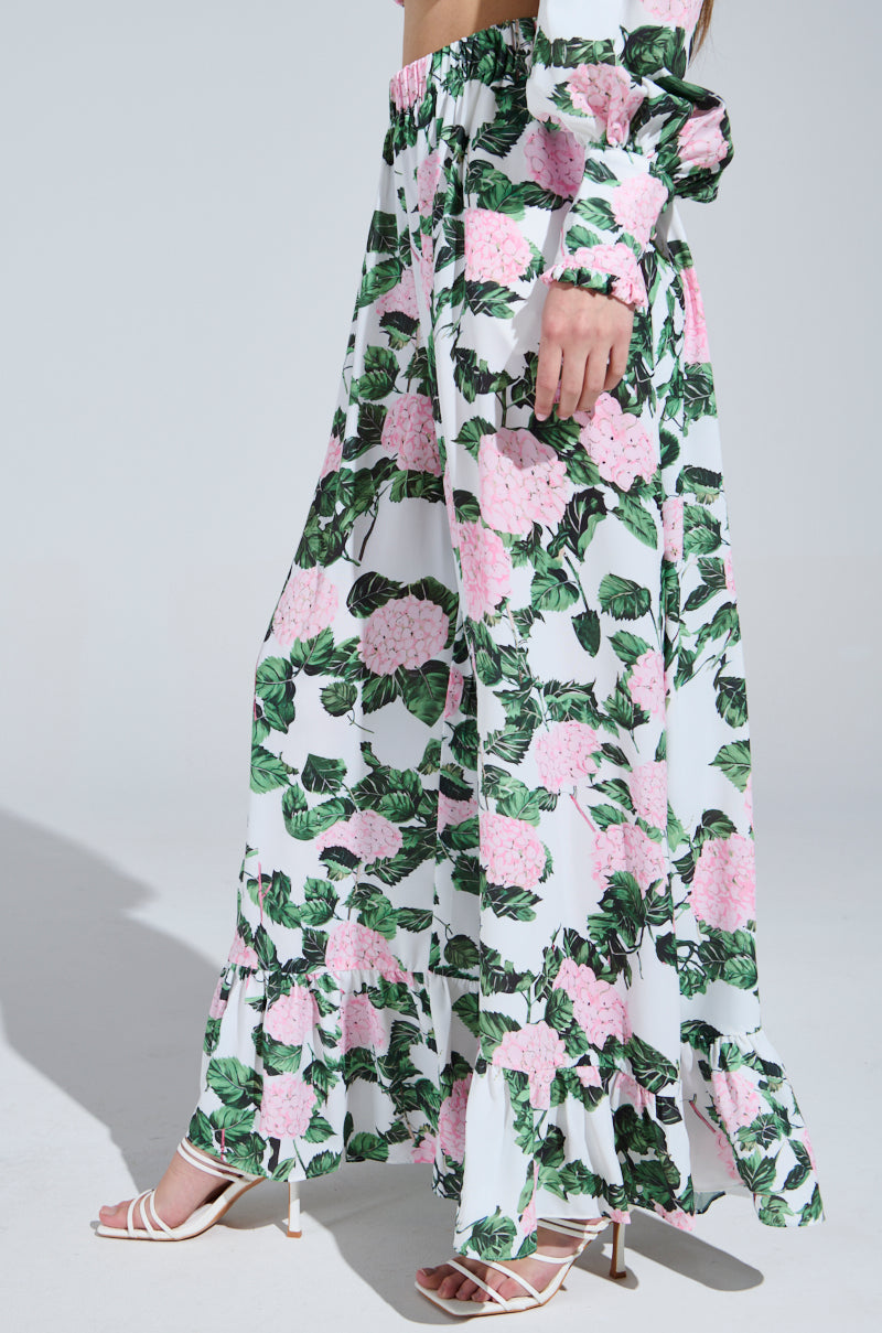 Someone wearing the DIANE FLORAL WIDE LEG RUFFLE PANT with a floral print of pink flowers and green leaves stands against a plain background. The flowing fabric complements their white strappy heels, with only part of their torso and legs visible.