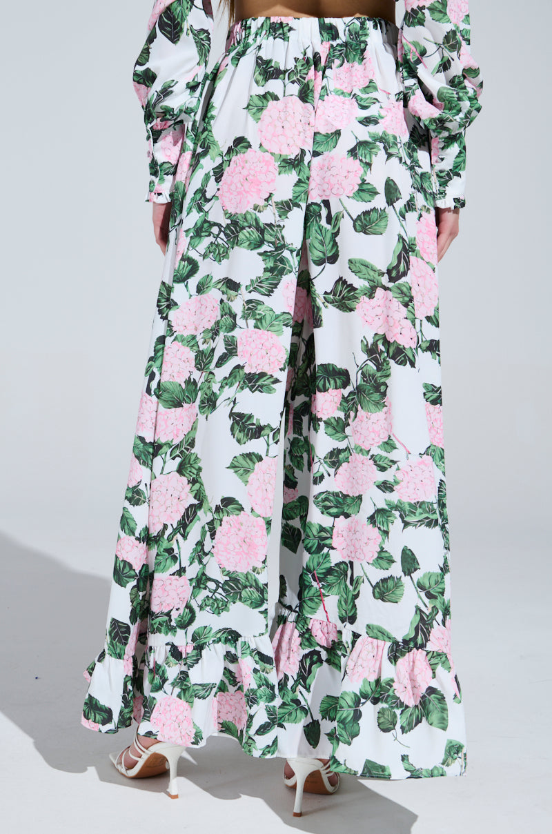 A person wears the DIANE FLORAL WIDE LEG RUFFLE PANT featuring a flowing satin design with pink flowers and green leaves, viewed from behind. The ruffled hem pairs beautifully with white open-toed shoes against a light, neutral background.