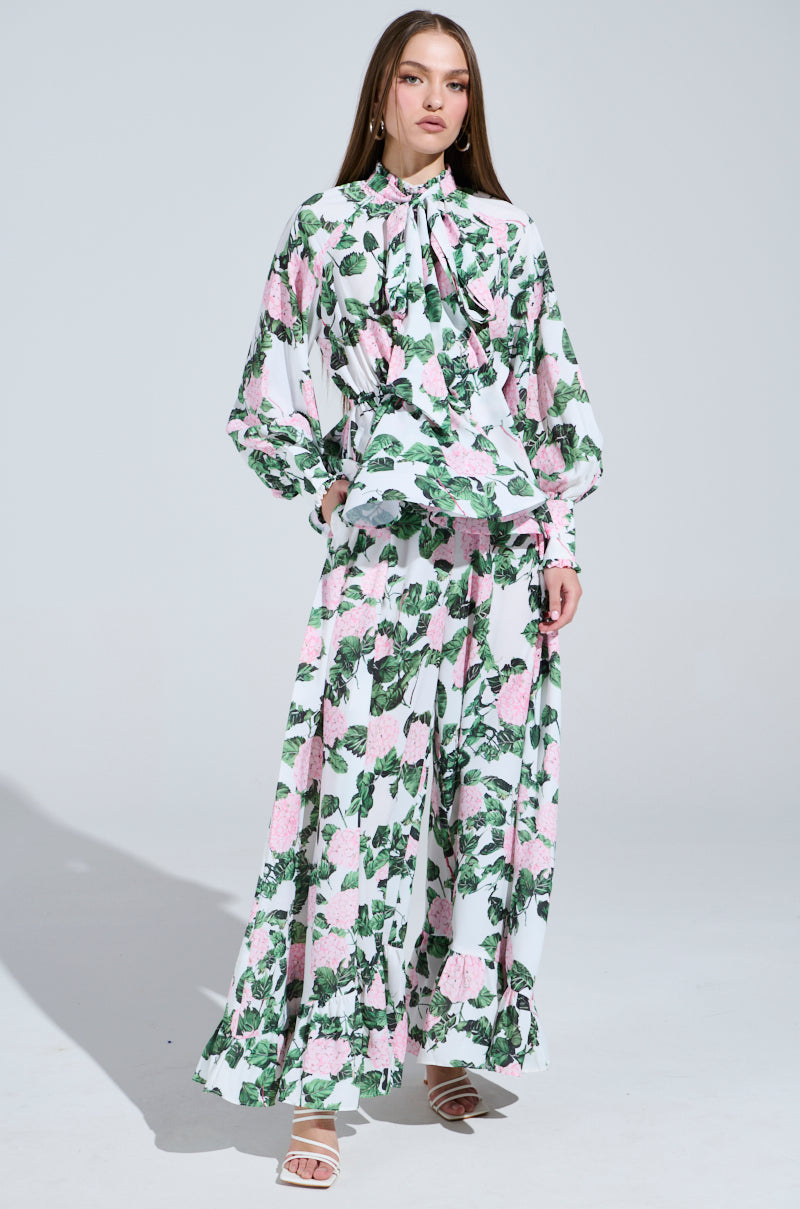A woman stands confidently in a flowing satin blouse and DIANE FLORAL WIDE LEG RUFFLE PANT, adorned with a pink and green pattern. Her white heeled sandals enhance her look, while her long straight hair frames her poised expression against a light background.