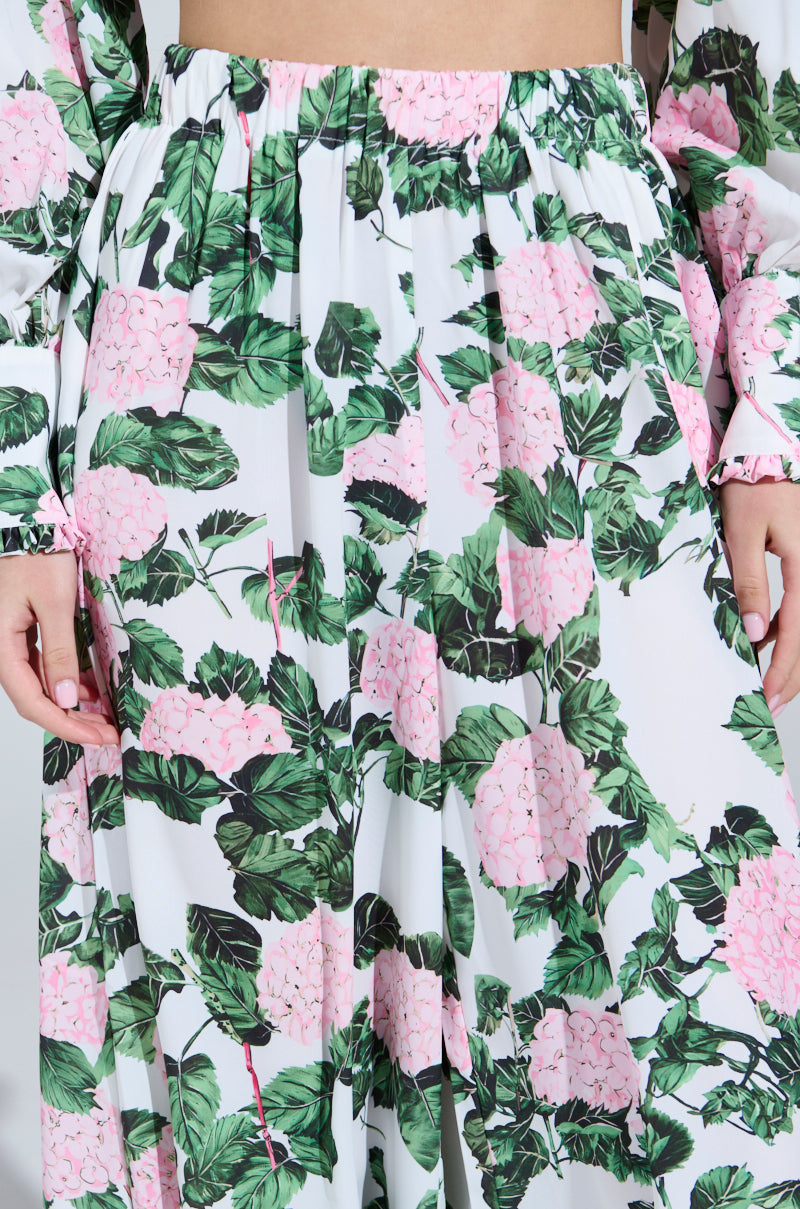 Wearing the DIANE FLORAL WIDE LEG RUFFLE PANT, showcasing pink hydrangeas and green leaves, a person stands out against the plain white background. The satin finish of the lower torso and hands enhances the ruffle pant style.