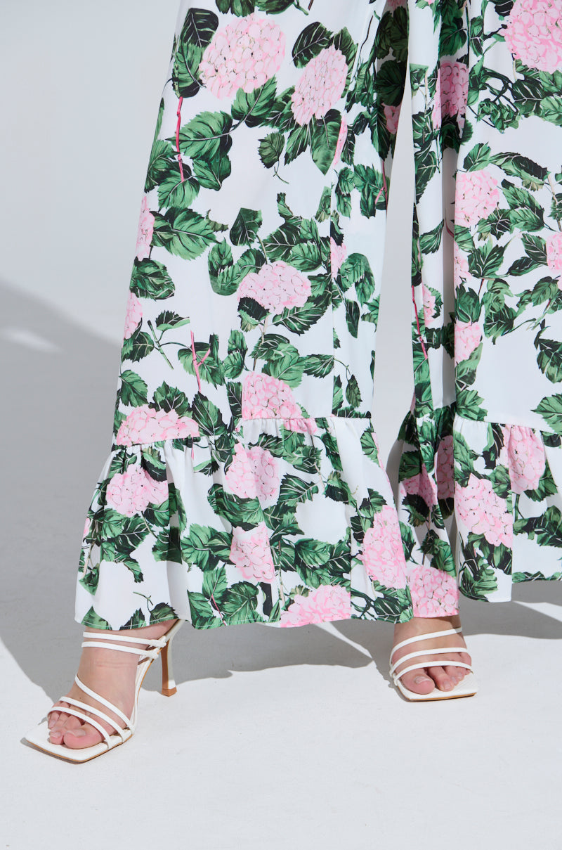 The DIANE FLORAL WIDE LEG RUFFLE PANT features a pink and green floral design on satin fabric with a ruffled hem, creating flair. It's elegantly paired with white strappy high-heeled sandals against a plain light background.