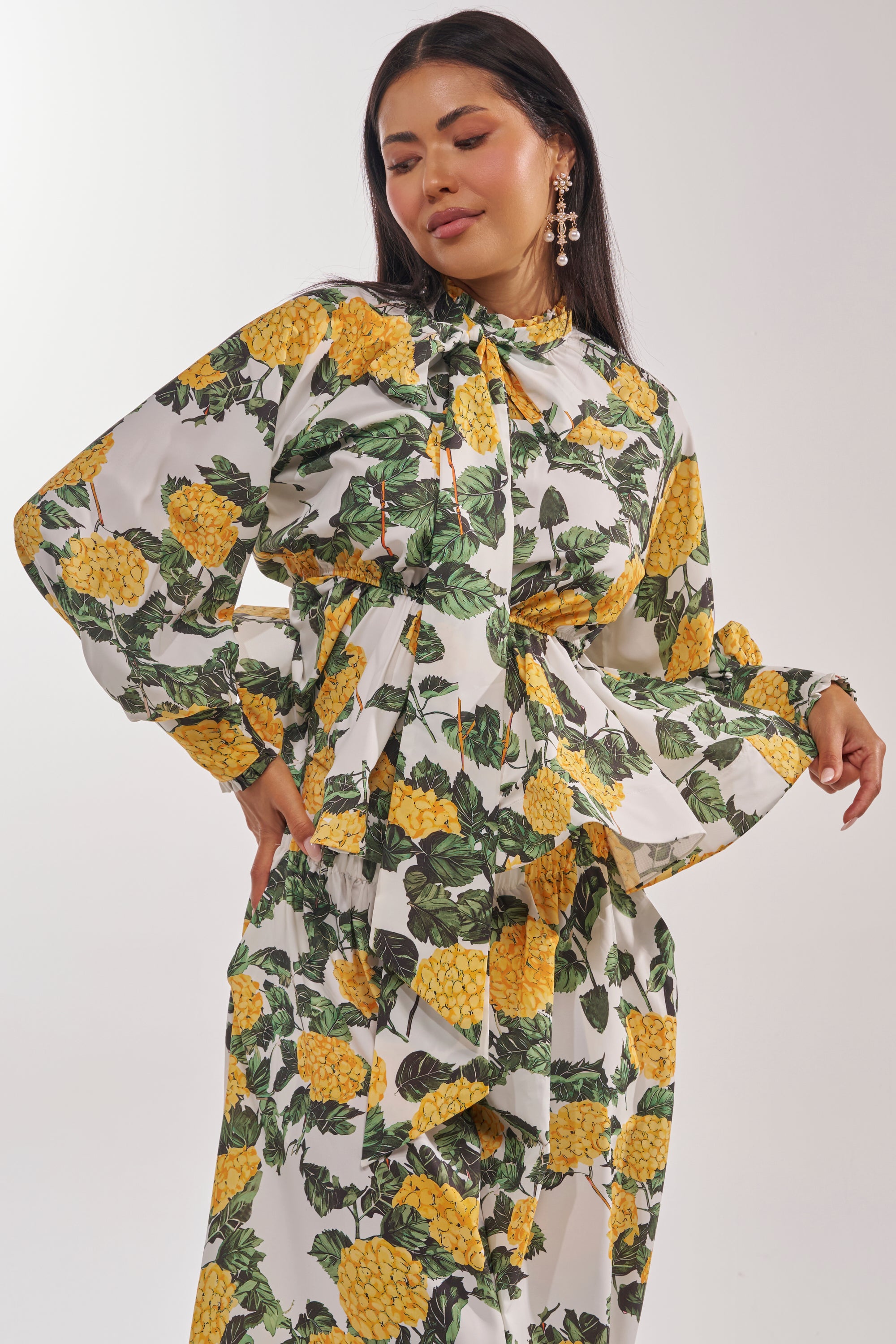 A woman with long dark hair wears the JULIE FLORAL PEPLUM BLOUSE, a yellow top featuring a green leafy print and long sleeves, paired with statement earrings. She stands against a plain white background. 1