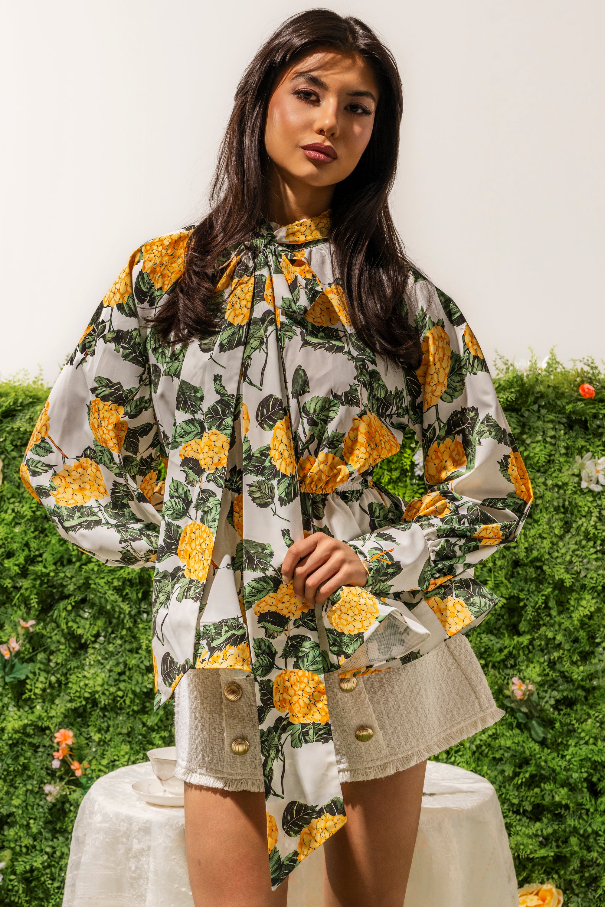 A woman with long dark hair models the JULIE FLORAL PEPLUM BLOUSE, featuring a yellow floral print and tie-neck bow, paired with white textured shorts with gold buttons, standing before lush green foliage for an effortless resort look. 0