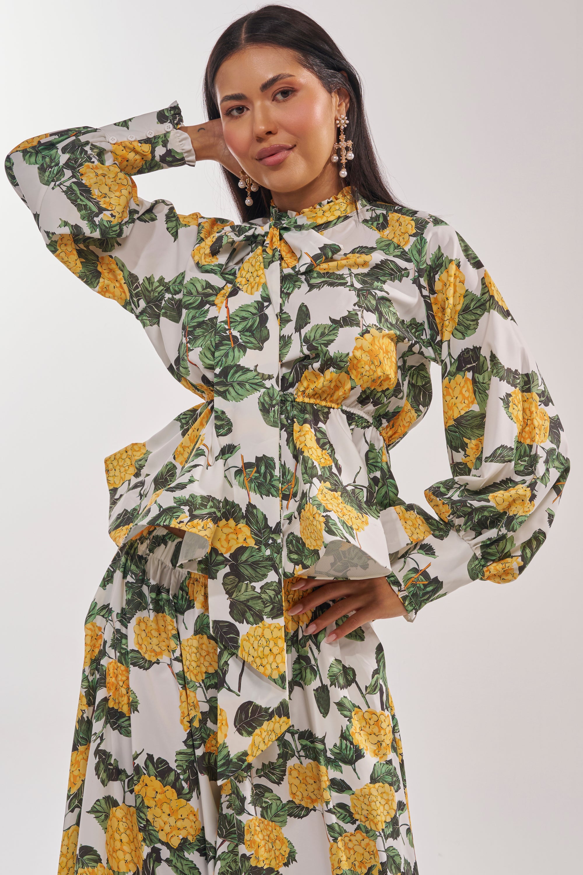 A woman poses confidently in the JULIE FLORAL PEPLUM BLOUSE, a high-neck yellow top with hydrangea and green leaf patterns, chic long sleeves, and statement earrings. She pairs it with a matching flared skirt. 2