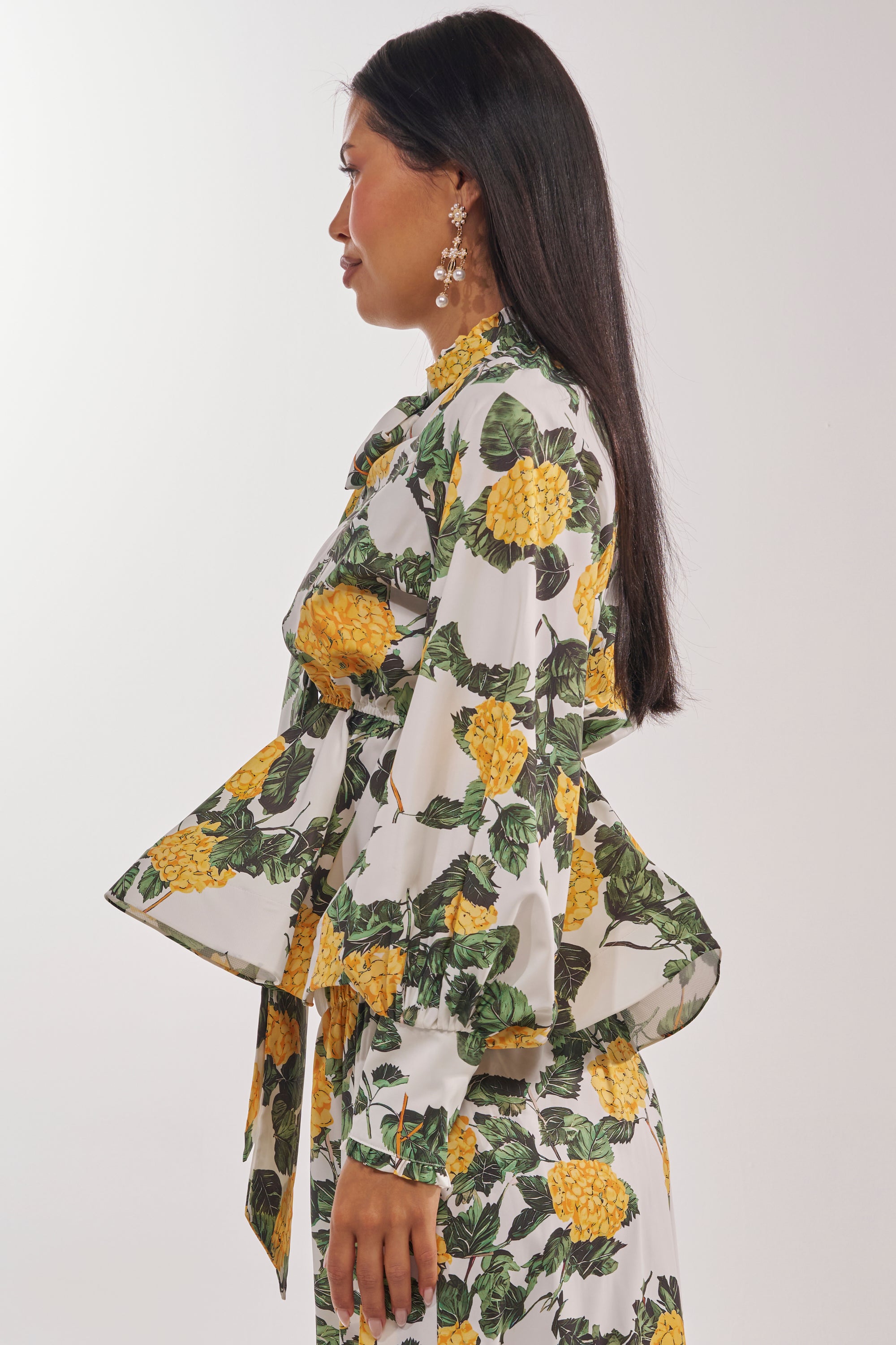 A woman with long dark hair stands in profile against a white background, wearing the JULIE FLORAL PEPLUM BLOUSE in yellow with green leaf prints, paired with a matching skirt and dangling floral earrings. 3