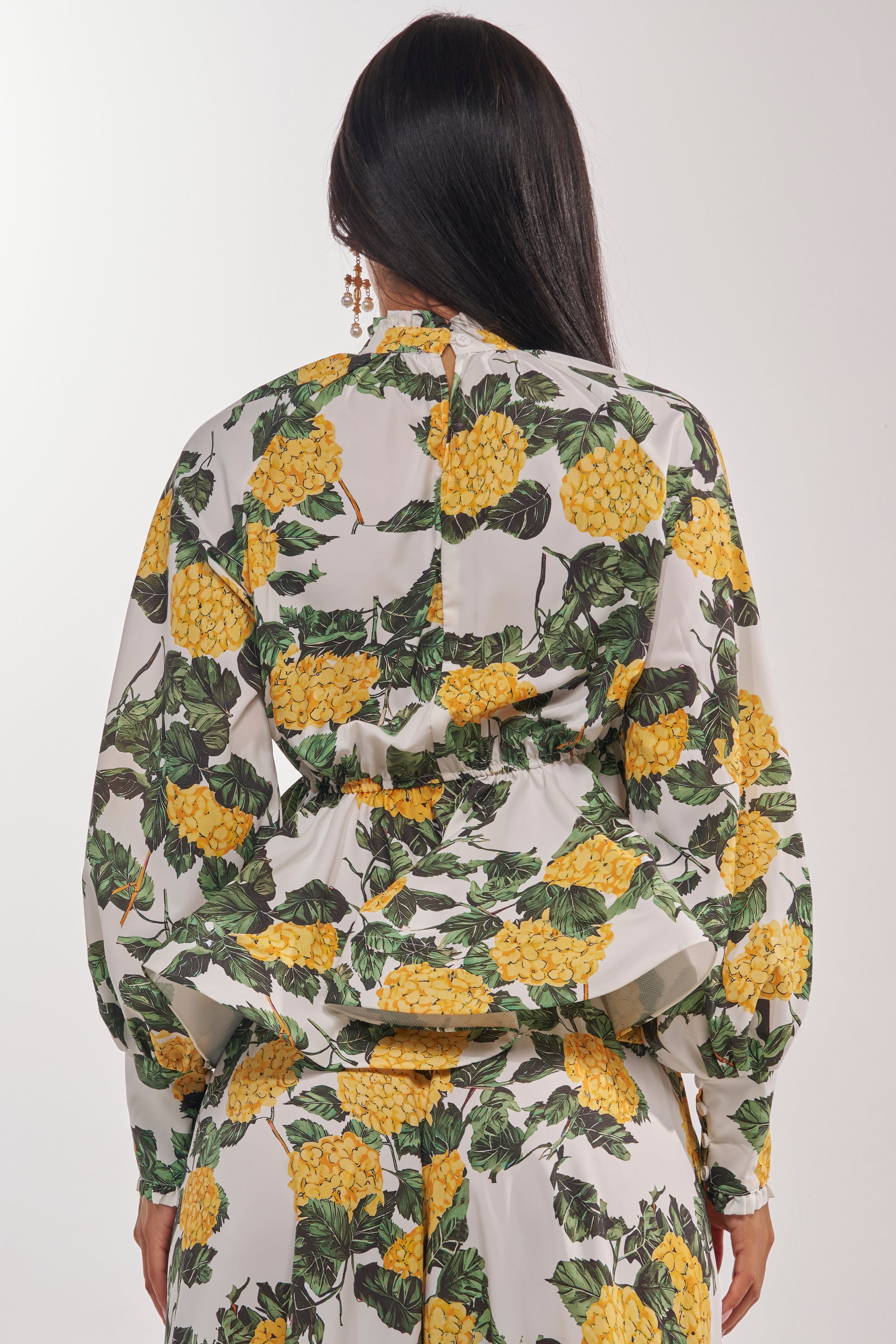 A woman with long, straight dark hair is seen from the back wearing the JULIE FLORAL PEPLUM BLOUSE, featuring a tie-neck detail and a floral pattern of large yellow flowers and green leaves on white—ideal for resort styling. 4