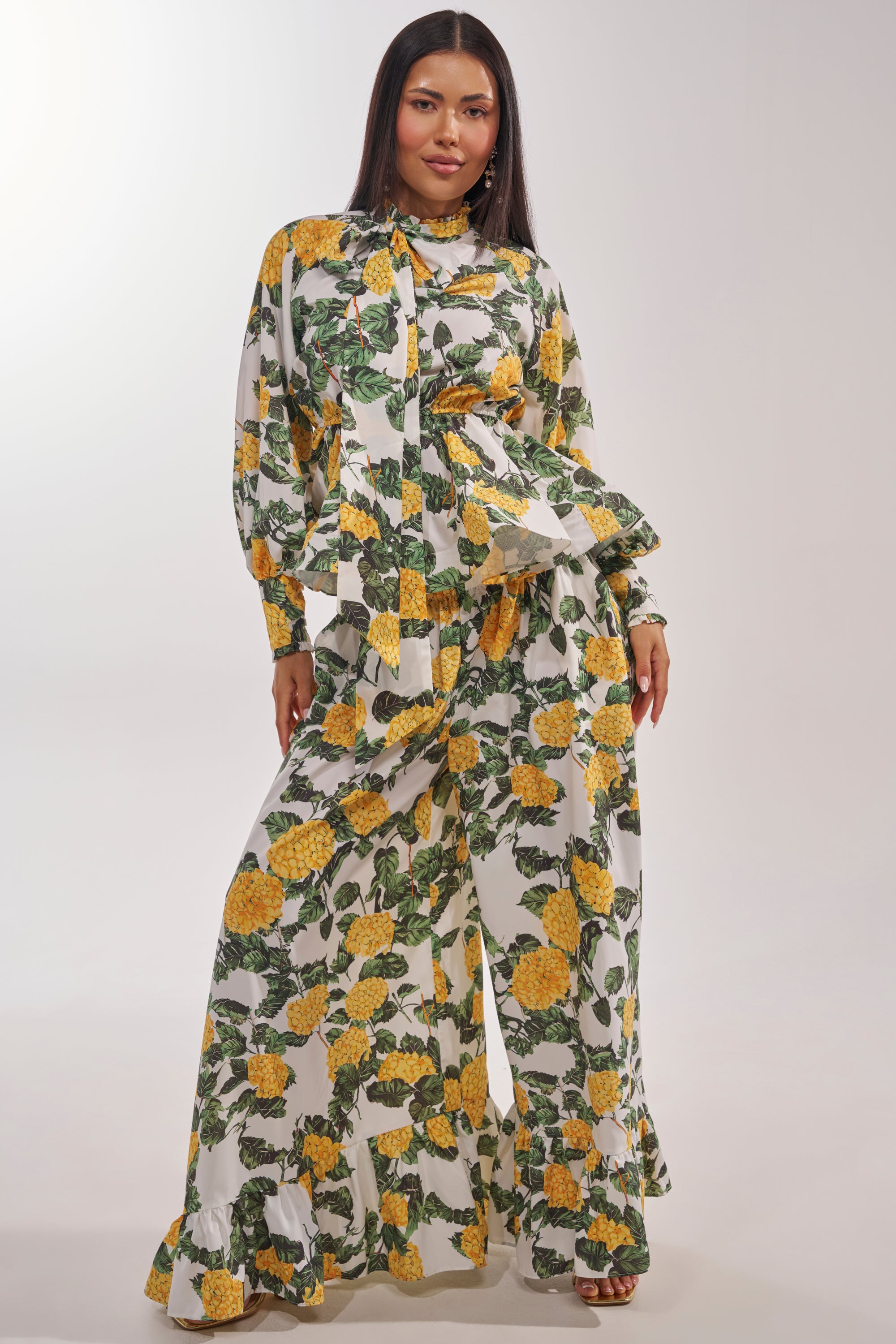 A woman exudes confidence in the JULIE FLORAL PEPLUM BLOUSE and wide-leg pants with yellow flowers and green leaves, capturing effortless resort style against a simple light background. 5
