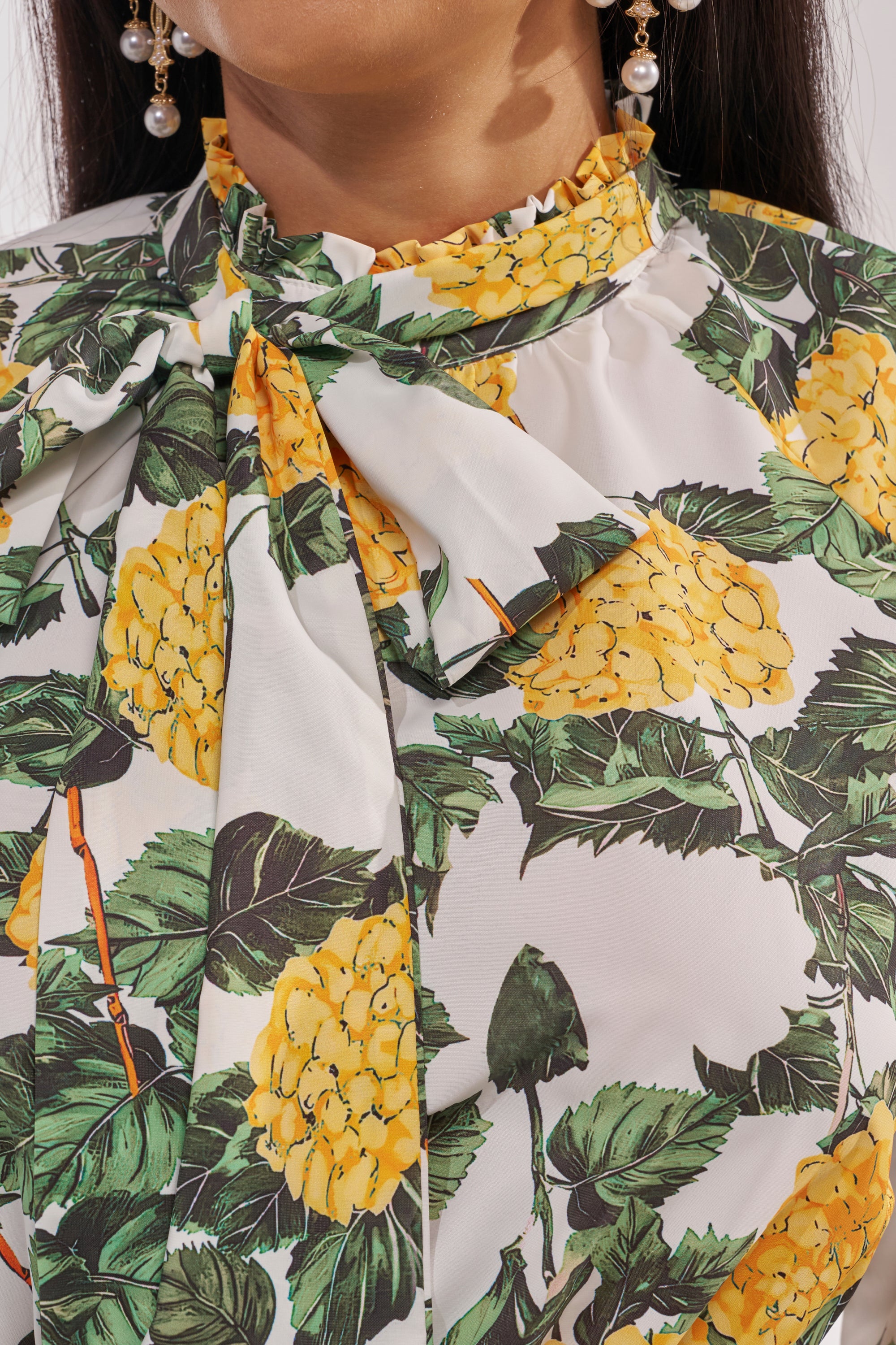 Close-up of a woman wearing the JULIE FLORAL PEPLUM BLOUSE, a yellow top with green leaf prints, tie-neck detail, and ruffled collar—perfect for resort looks. She completes the style with dangling pearl earrings. 7