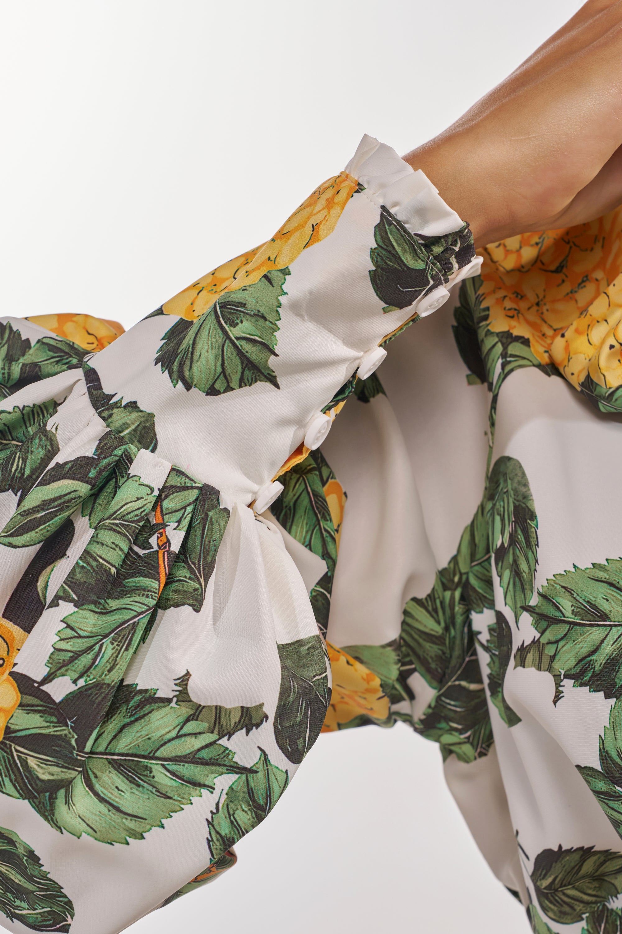 A close-up of an arm wearing the JULIE FLORAL PEPLUM BLOUSE—yellow with green leaf print, puffy sleeves, and fabric-covered buttons—perfect for fresh resort style. 8