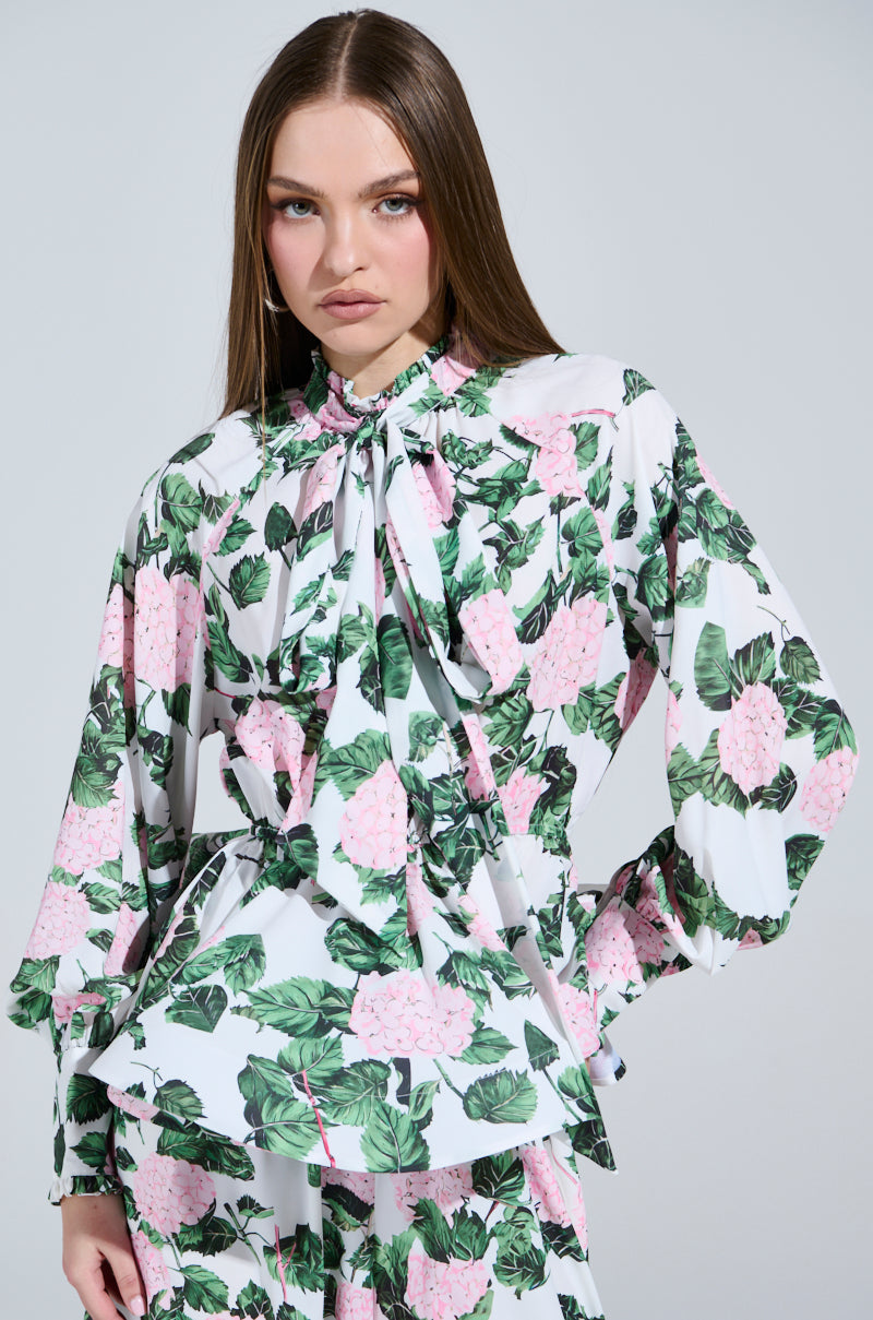 A person with long brown hair wears the DIANE FLORAL PEPLUM BLOUSE, showcasing a chic floral design with pink flowers and green leaves on white. It features a neck tie and ruffled details, set against a light gray background, wearing a neutral expression.