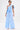 A person poses in the light blue ASHLEY RELAXED PANT, featuring a high-rise wide-leg silhouette and subtle striped cotton-blend fabric. Styled with a white clutch, white shoes, slicked-back hair, statement earrings, and a plain white background.