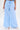 A person stands against a white background wearing the light blue ASHLEY RELAXED PANT in a high-rise, wide-leg fit, paired with white open-toe heels and a small white handbag.
