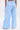 A model is seen from the back wearing the ASHLEY RELAXED PANT, featuring loose-fitting, light blue and white pin stripes. The high-rise, wide-leg pants are styled with a white handbag and heeled shoes against a plain background.