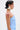 A woman with short, slicked-back hair and a white floral earring models the ASHLEY BRALETTE, a light blue, sleeveless, ruffled cotton-blend top. She poses against a plain light background with a bold red abstract design on her upper arm and shoulder.