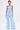 A woman stands against a white background wearing the ASHLEY BRALETTE, a light blue strapless smocked top, with matching wide-leg pants. She accessorizes with a white handbag, white heels, drop earrings, and displays visible tattoos.