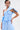 A person with short dark hair and statement earrings wears the ASHLEY BRALETTE, a light blue, asymmetrical off-shoulder top with pinstripes, paired with matching loose pants. They have an arm tattoo and a neutral expression.