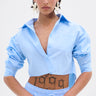 A person with short dark hair wears statement earrings and the ASHLEY BUTTON DOWN TOP with matching pinstripe pants. They stand confidently, revealing visible tattoos, including "1999" on their torso.