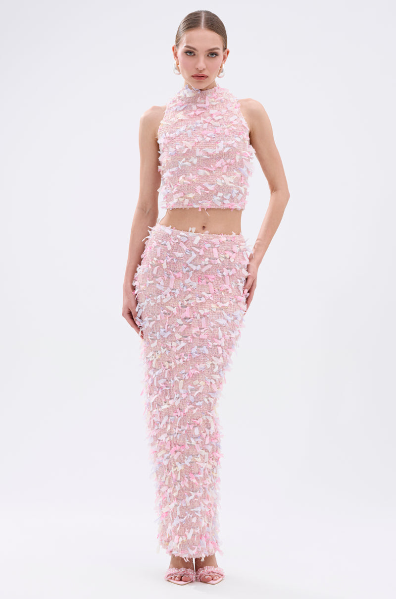 A woman models the WILD AND FREE FRINGE MAXI SKIRT—a sleeveless, high-neck pale pink crop top and matching maxi skirt set with pastel feather-like embellishments and fringe detail. She pairs it with pink heels against a plain white background.