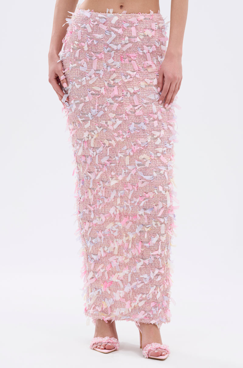 A person wears the WILD AND FREE FRINGE MAXI SKIRT, a long, fitted pink skirt featuring textured multicolored fringe, paired with matching pink open-toe heels. The high-rise fit and pastel hues add decorative, tactile appeal.