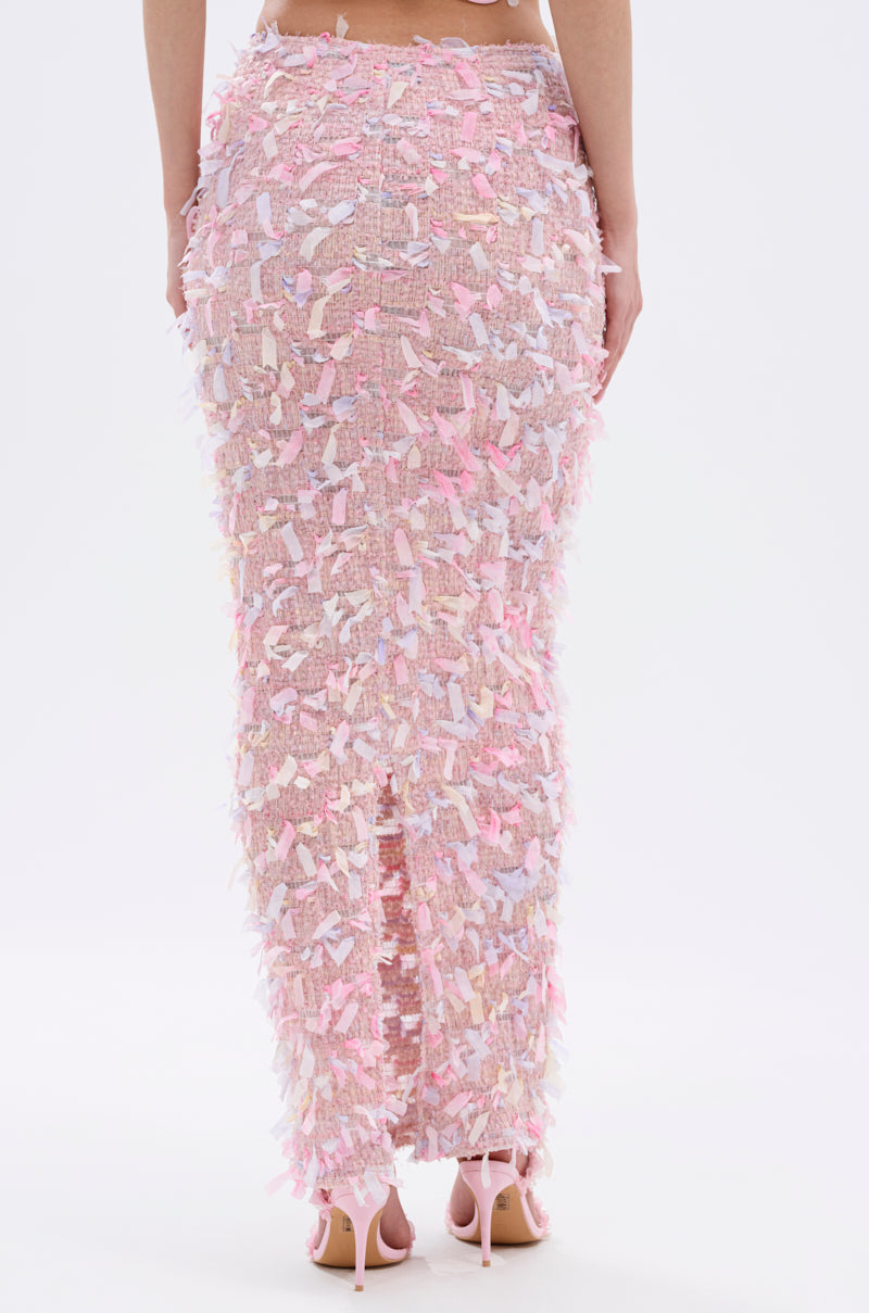 A person wears the WILD AND FREE FRINGE MAXI SKIRT, a long fitted pink mesh skirt with pastel details and a subtle back slit, viewed from behind. It's styled with light pink high-heeled shoes.