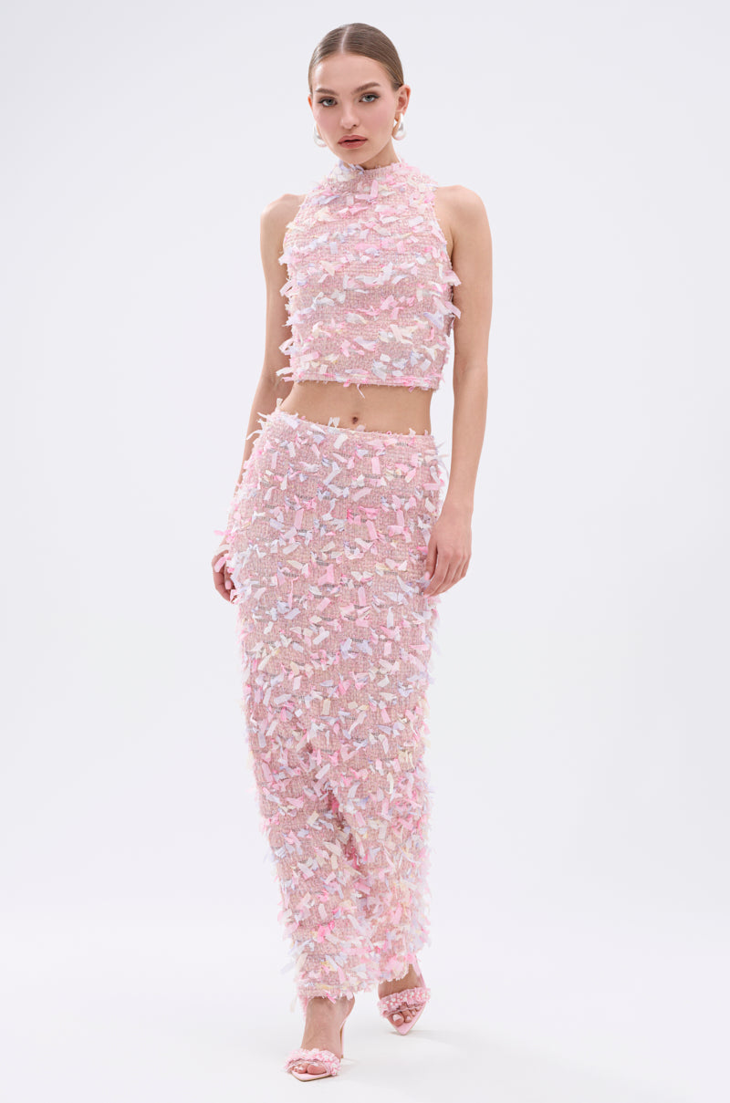 A woman models the WILD AND FREE FRINGE MAXI SKIRT, a pastel pink, white, and lavender maxi skirt with fringe detailing, paired with a matching sleeveless cropped top, open-toe pink heels, and her hair pulled back.