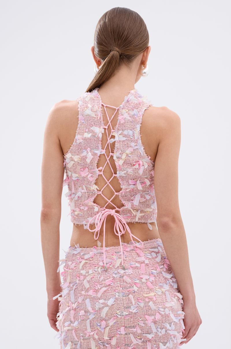 A woman faces away, wearing the WILD AND FREE FRINGE BLOUSE—a sleeveless pastel pink crop top with a crisscross lace-up back and pastel fringe—paired with a matching high-waisted skirt. Her hair is in a low, sleek ponytail.
