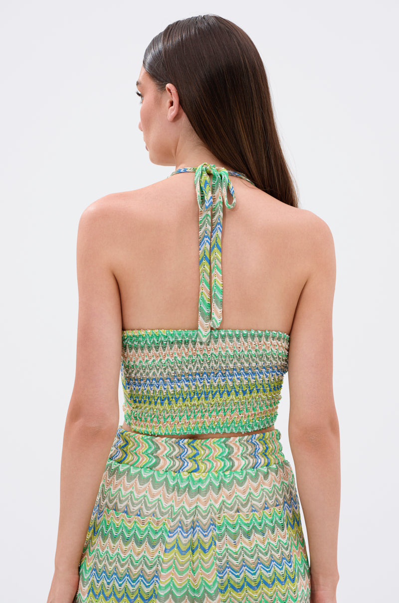 A woman with straight brown hair faces away from the camera, wearing the JUST A FEELING CROCHET HALTER TOP in green, blue, and white with a zig-zag chevron pattern, open back, smocked bodice, and halter neck ties.