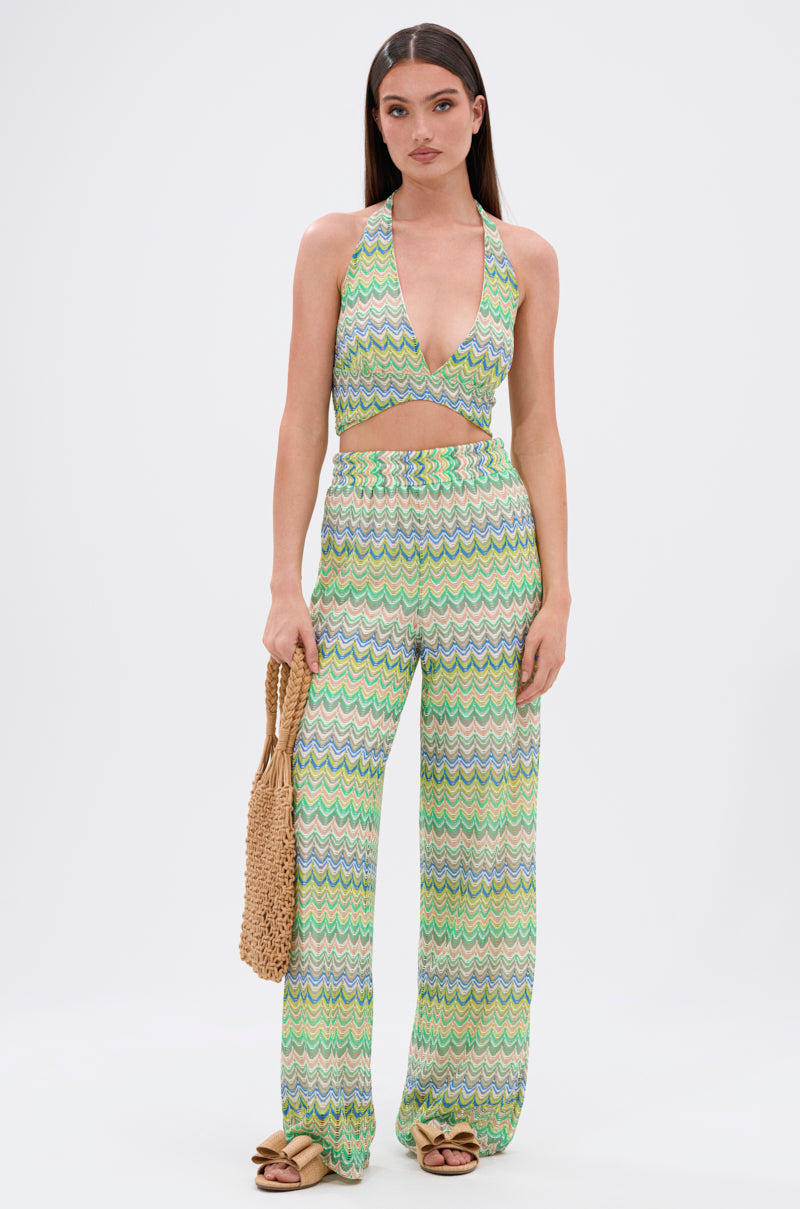 A woman stands against a plain background wearing the JUST A FEELING CROCHET HALTER TOP in a green, yellow, and white chevron pattern with matching high-waisted, wide-leg pants, holding a woven bag and wearing light brown sandals.