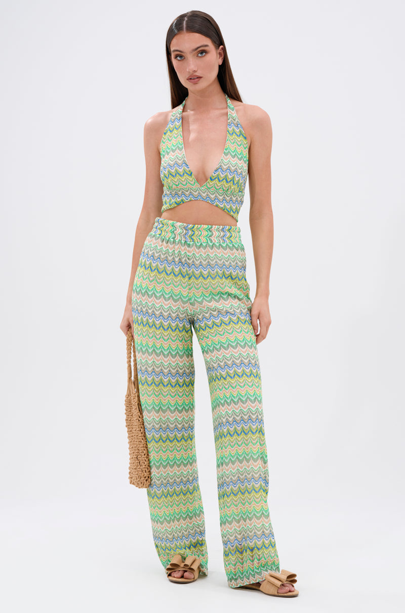 A woman faces forward in the JUST A FEELING CROCHET HALTER TOP, a sleeveless green and blue chevron crochet halter with open back, paired with matching high-waisted pants. She holds a woven bag and wears tan sandals against a white background.