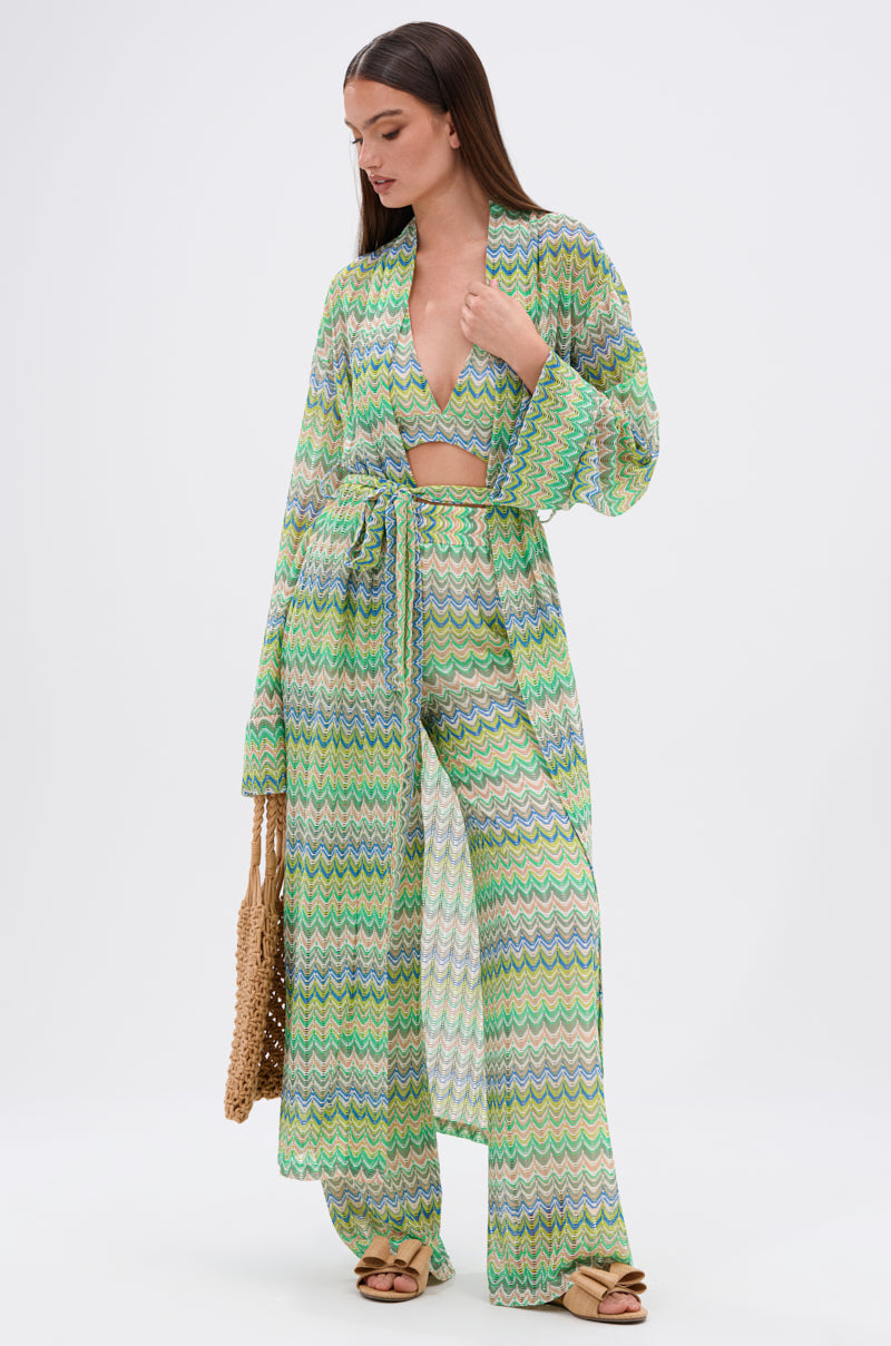A woman poses against a white background wearing the JUST A FEELING CROCHET HALTER TOP in green, blue, and white chevron pattern, paired with wide-leg pants, a long robe, tan sandals, and holding a woven bag.