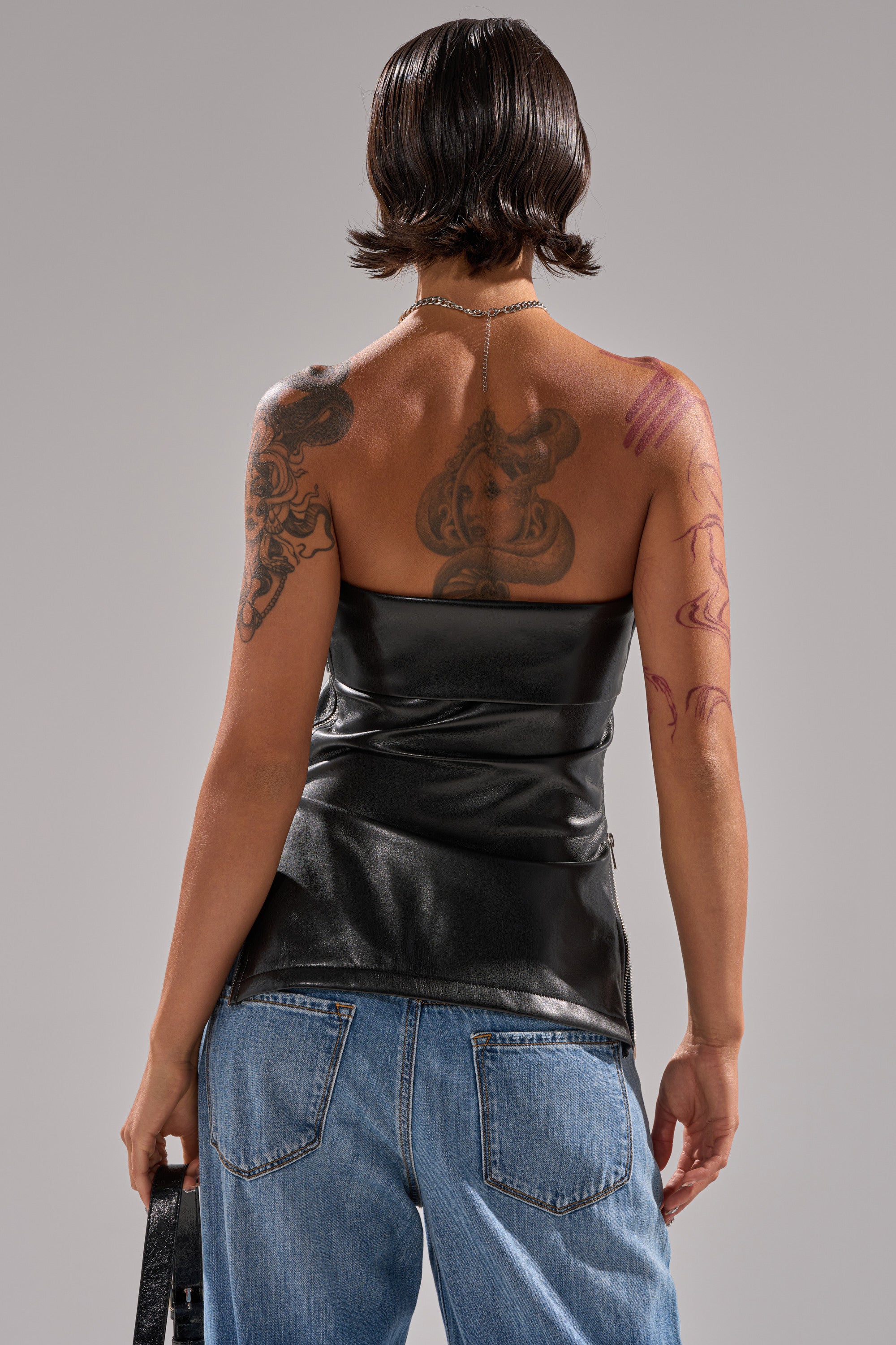 A person with short, wet-look hair is seen from behind, wearing the black ZIP ME UP ON THE SIDE CORSET strapless top and blue jeans. Their arms and back have multiple tattoos, and they hold a black bag in their left hand. 4