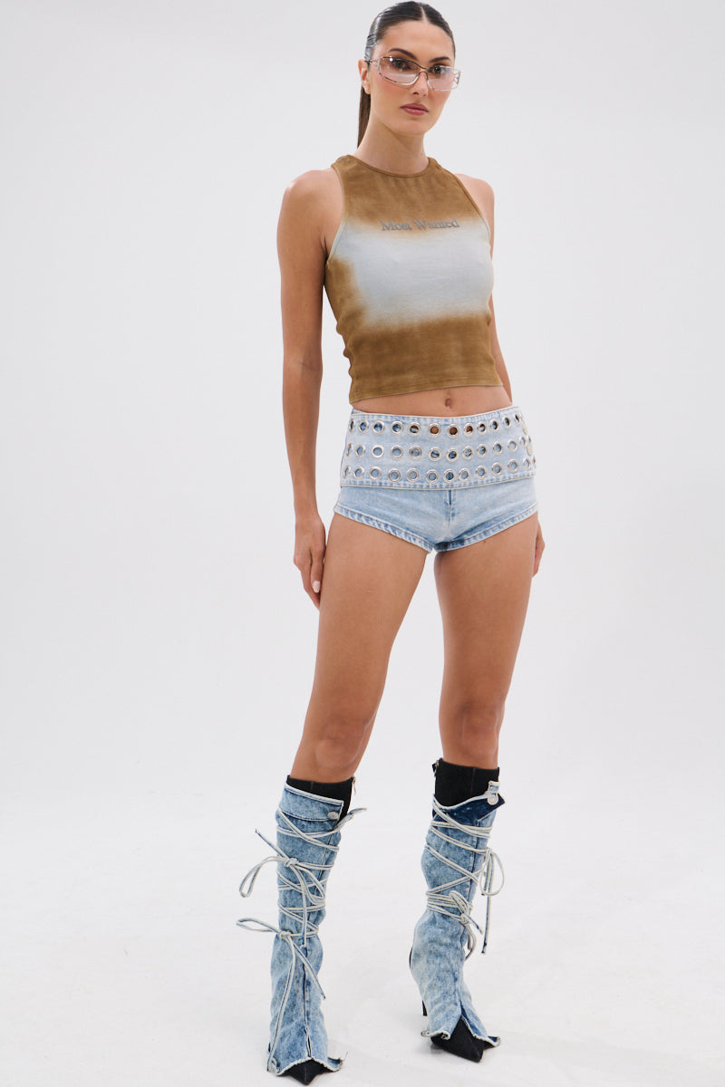 A woman stands against a white background wearing the MOST WANTED TANK, a brown and white ribbed cotton-blend sleeveless crop top, paired with high-waisted denim shorts with silver grommets and lace-up acid-wash denim boots.