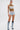 A woman stands against a white background wearing the MOST WANTED TANK, a brown and white ribbed cotton-blend sleeveless crop top, paired with high-waisted denim shorts with silver grommets and lace-up acid-wash denim boots.